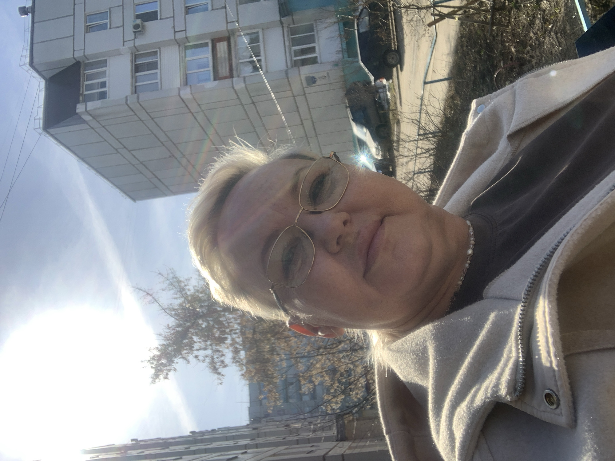A woman with short blonde hair and glasses is taking a selfie outdoors. She is wearing a brown shirt underneath a light-colored jacket. Behind her is a building with multiple windows, seemingly located in an urban area. The sun is shining brightly, creating glare. Parked cars are visible in the background, along with bare trees suggesting it may be autumn or winter. Considering the location in Chaikovsky, Russia, based on the other context provided, the building's architecture appears typical of Soviet-era construction. The overall scene suggests a casual, everyday moment captured in an urban environment.