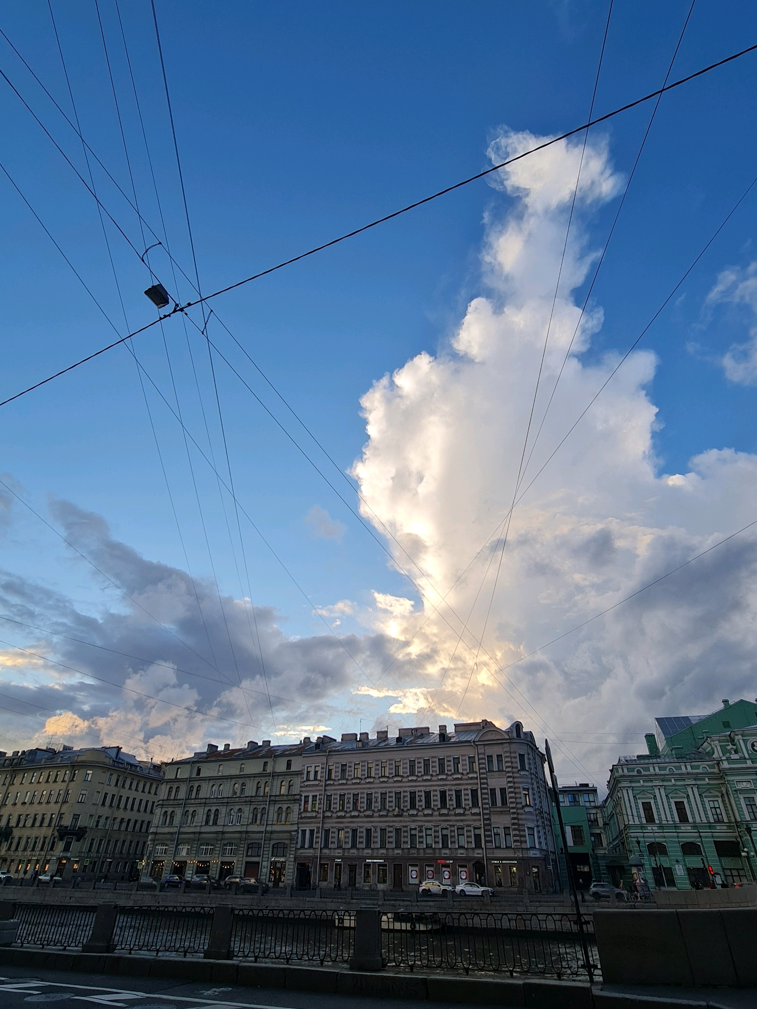 This is a street-level, wide-angle photograph of a canal-side cityscape in Saint Petersburg, Russia, likely taken during the daytime given the bright sky. The foreground features a dark, narrow canal bordered by a metal railing and a paved walkway. Beyond the canal, a row of multi-story buildings with ornate classical architecture lines the embankment. The buildings vary in color and architectural detail, with some exhibiting pastel hues and others a darker, more muted palette. Visible in the street behind the canal are several parked cars, suggesting urban activity. Above the buildings, a dramatic sky dominates the scene, filled with a mix of bright blue expanses and large, billowing white clouds, some of which are illuminated by sunlight. A prominent network of dark utility wires crisscrosses the sky, creating a graphic element that intersects with the clouds and buildings. One street lamp is visible, suspended from the wires. The overall impression is of a historic European city, characterized by grand architecture and the presence of modern infrastructure.