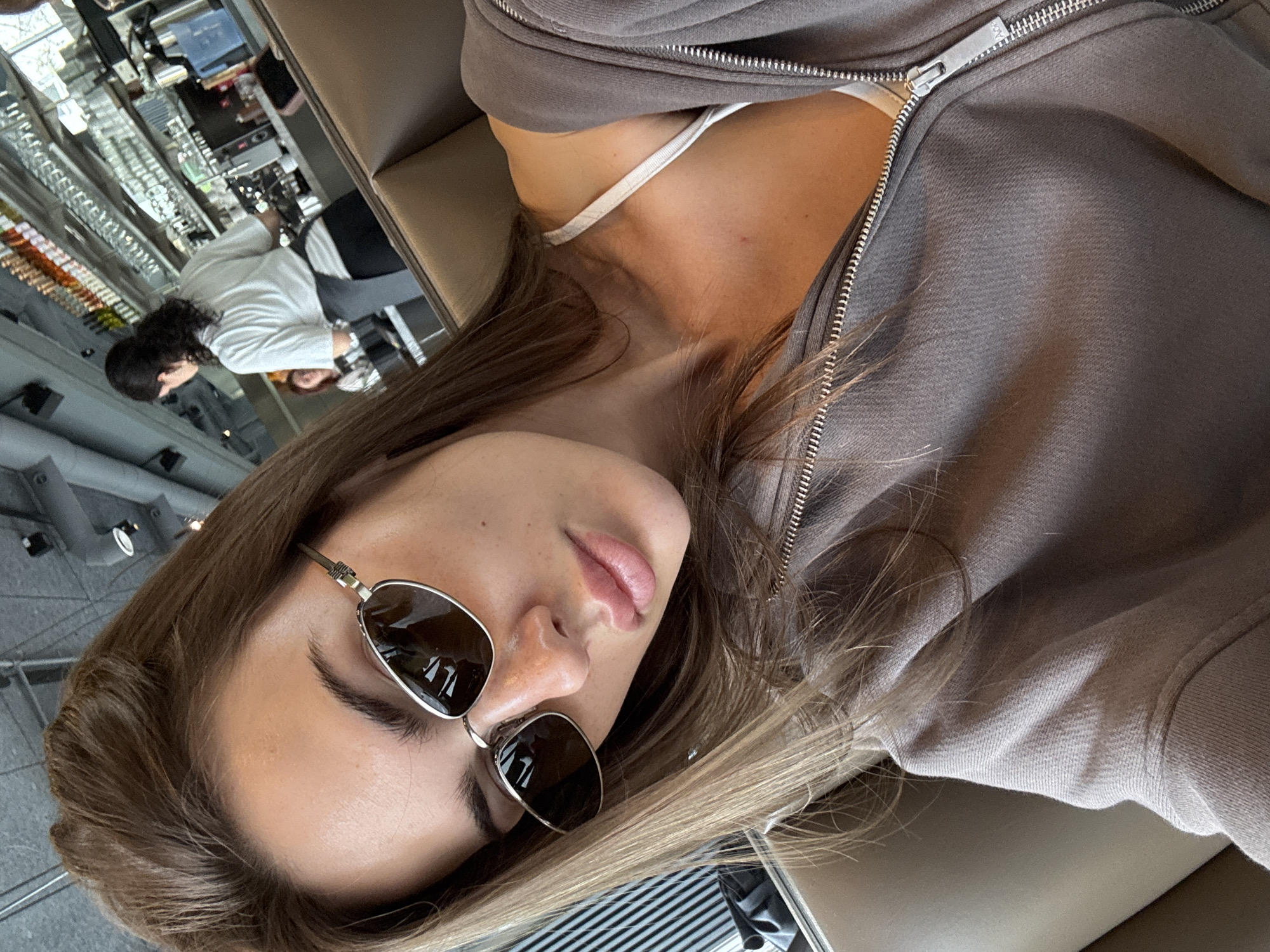 A woman wearing sunglasses and a partially unzipped grey sweatshirt is lying down. A thin white strap of fabric is visible under the sweatshirt. In the blurry background, other people are present in what appears to be a restaurant or cafe. The reflection in the sunglasses shows objects on the ceiling that look like lights or vents. Based on these details, it is difficult to definitively determine the location within Warsaw.