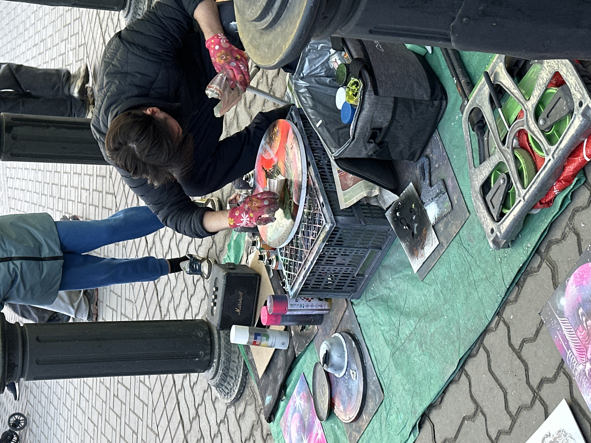 An outdoor, daytime scene in a paved urban pedestrian area, identified as being in Kazan, Russia, depicts a street artist actively creating spray paint art. A person with dark hair, wearing a black puffer jacket, black pants, and pink patterned gloves, is crouched over a circular artwork, manipulating paint with their gloved right hand and holding a crumpled material in their left. The artwork in progress displays vibrant reds, oranges, and purples, depicting a cosmic or stylized landscape, and rests on a dark plastic crate.
The artist's workspace is established on a crumpled green tarp laid over grey interlocking concrete pavers, which feature a wavy design. Surrounding the artist are numerous tools and supplies: a black Marshall portable speaker, multiple spray paint cans (including one white can with "RALIN" visible and two reddish-pink cans), a grey soft-sided cooler bag containing additional paint cans and containers, various stencils, a small metal bowl, a metal plate, and other metal tools. Several completed or partially completed flat artworks with colorful, cosmic themes are visible on the left side of the tarp.
In the background, partially obscured by dark, fluted bollards, are three other individuals. One person is standing upright, wearing a light blue puffer jacket, blue sweatpants, and light-colored sneakers. The lower legs and feet of two other people are visible further in the background, indicating the presence of other pedestrians in the public space. The scene is illuminated by natural daylight.