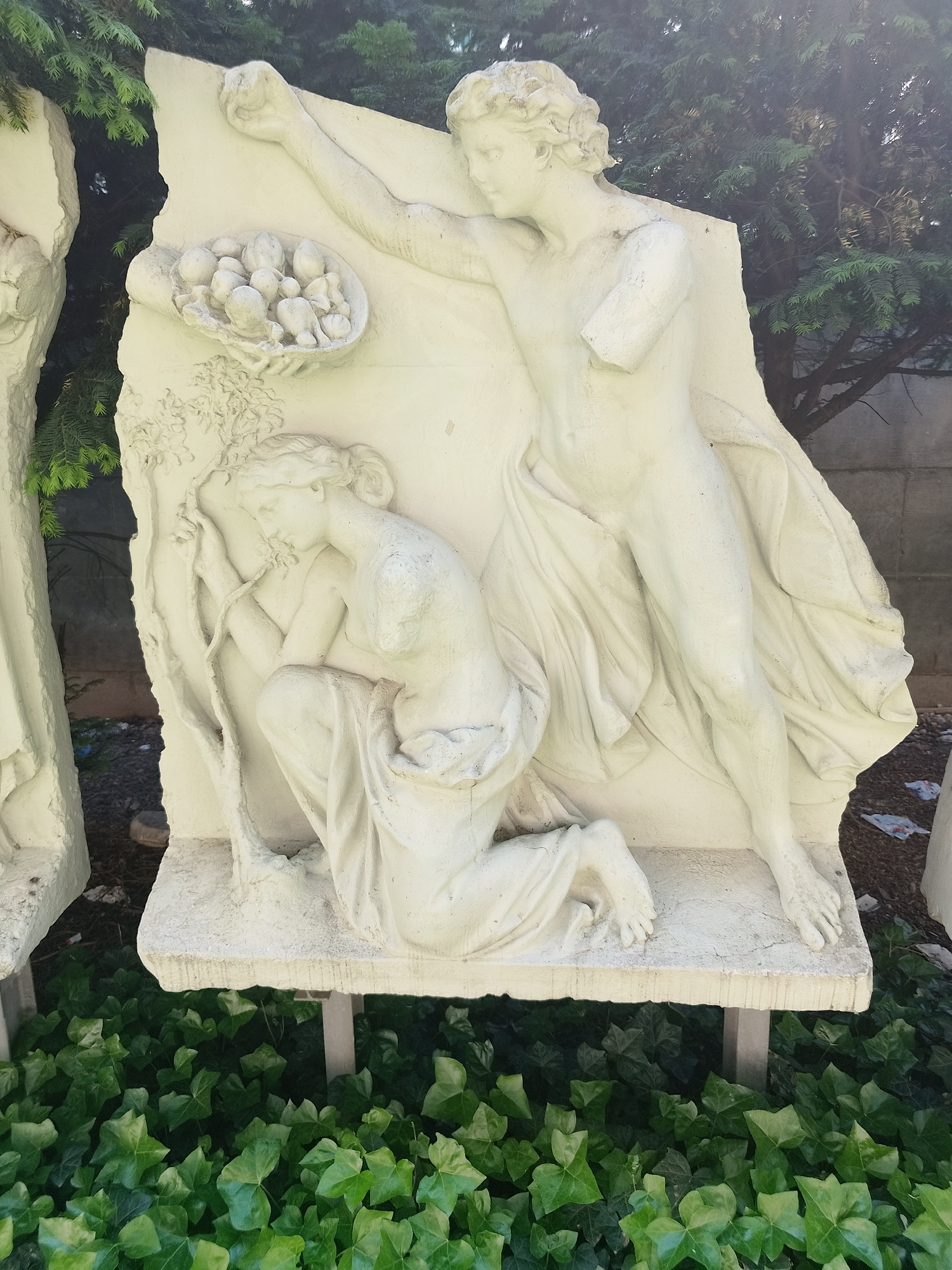 A photograph captures a white concrete relief sculpture featuring two classical figures. One figure stands, missing part of an arm, draped in flowing fabric, with an arm raised holding a dish of fruit. A second figure kneels near a tree, partly obscured by the standing figure. The sculpture rests on a concrete base among ground covering greenery. The exact location within the city of Unknown, Unknown is not discernible from the photograph.
