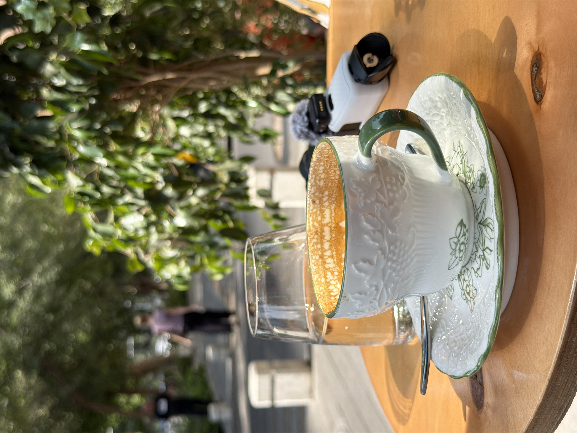 A close-up, eye-level shot captures a coffee cup and a glass of amber liquid on a wooden table. The coffee cup, white with intricate embossed patterns and a green handle, sits on a matching saucer adorned with green floral designs. The coffee inside has a foamy, light brown surface. To the left of the cup, a clear glass filled with the amber liquid stands tall.
In the background, blurred figures of people walk along a paved pathway. Lush green trees with dappled sunlight filtering through their leaves create a vibrant, natural backdrop. The scene appears to be set outdoors, possibly in a park or an outdoor cafe area. The overall ambiance is relaxed and leisurely. This setting is consistent with the outdoor seating areas found in popular cafes along Amir Timur Street in Tashkent, Uzbekistan, offering a pleasant urban green space.