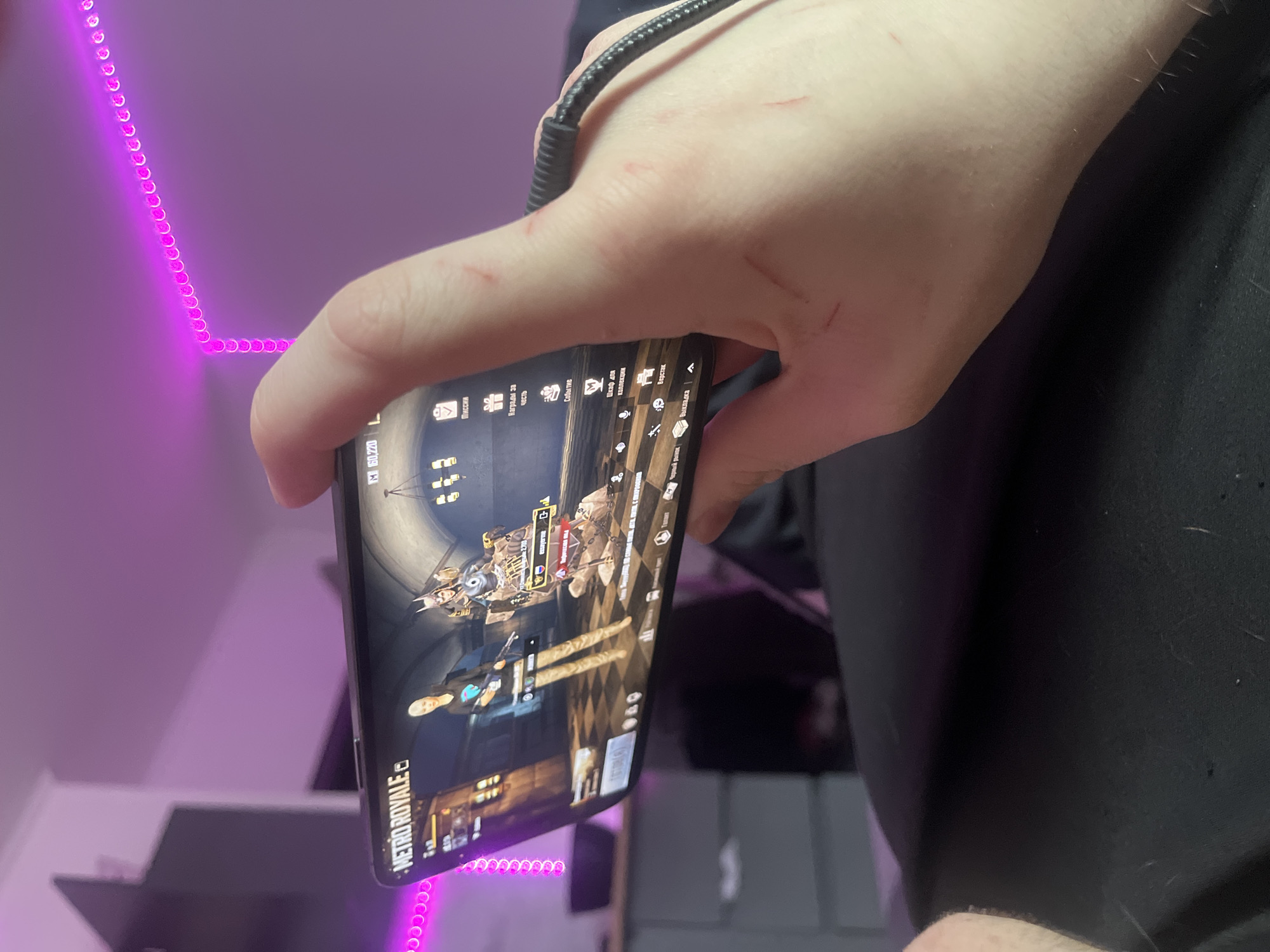 An overhead, portrait-oriented shot captures a human hand holding a black smartphone, actively displaying a mobile game. The phone screen shows an in-game menu with character models and various options, predominantly in Russian. Visible text includes the game title "METRO ROYALE," and menu options such as "Миссии" (Missions), "Награды за честь" (Honor Rewards), "Событие" (Event), "Шкаф для коллекции" (Collection Cabinet), "Черный рынок" (Black Market), "Верстак" (Workbench), and "Выкладка" (Layout/Display), along with "160,220 L."
A braided black USB-C cable is plugged into the top of the phone, running upwards out of frame. The back of the hand holding the phone has several small, faint red marks or scratches. The setting is indoors, likely a room, characterized by a vibrant L-shaped strip of pink/purple LED lights illuminating a wall in the background, suggesting an evening or nighttime ambiance. Other dark, out-of-focus objects, possibly furniture, are visible in the lower background. The presence of Russian text on the screen is consistent with the provided location context of Saint Petersburg, Russia, where a person is engaged in mobile gaming.