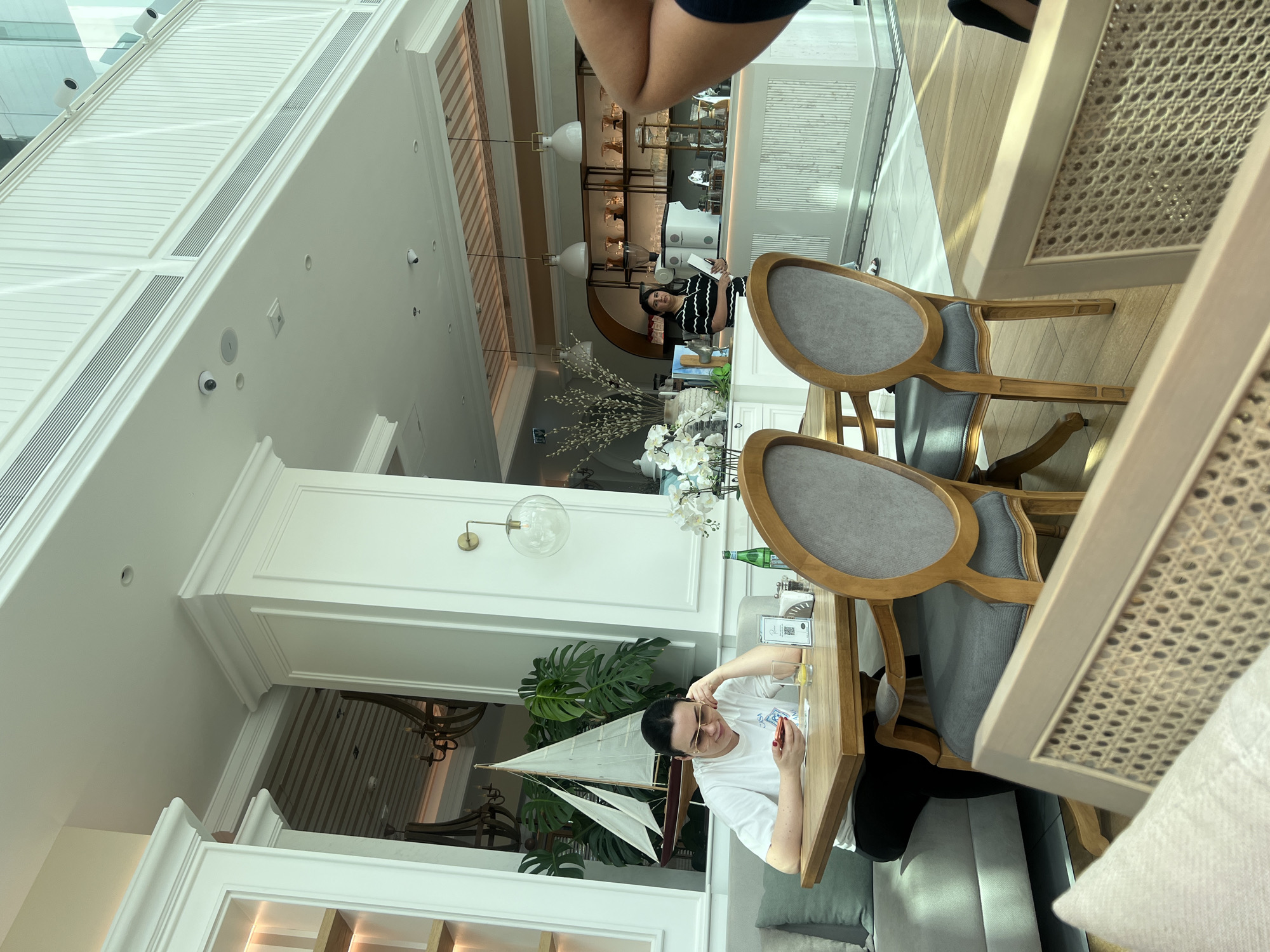 This image captures the interior of a spacious and elegantly decorated cafe or restaurant, likely located in Dubai, United Arab Emirates. The setting features high ceilings, light-colored walls with intricate moldings, and large windows or skylights that provide ample natural light, suggesting it is daytime.
In the mid-ground, a woman with dark hair, wearing a white t-shirt and dark trousers, is seated at a light wooden table. She is looking towards the camera with a neutral expression, wearing gold-rimmed glasses. She holds a small, rectangular object, possibly a mobile phone or a card. On her table, there is a glass containing a yellow beverage with a lemon slice, a bottle of sparkling water (identifiable as Pellegrino by its shape), and a small white sticker with a visible QR code. The seating around her includes a grey upholstered banquette and classic-style wooden chairs with grey fabric seats and cane backs.
In the background, further into the establishment, another woman in a black and white striped top is visible behind a service counter, possibly an employee. The counter area features glassware and shelves. Decorative elements throughout the space include a large model sailboat, potted green plants, and white artificial flowers, adding to the sophisticated ambiance. Ornate wall-mounted light fixtures and modern ceiling vents are also prominent. Several small security cameras are mounted on the walls and ceiling, indicating a monitored environment. The overall impression is one of a modern, upscale dining or lounge area.