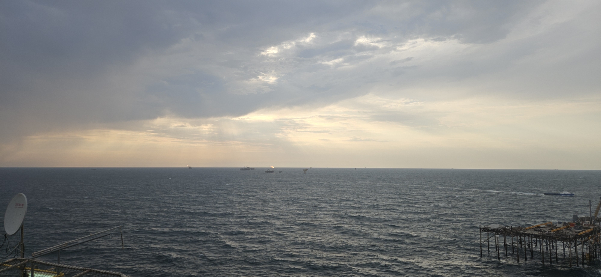 This is a wide shot of an offshore platform or an oil rig at sea, with a vast expanse of dark, choppy water under a cloudy sky. Sunlight is breaking through the clouds in some areas, creating beams of light. In the foreground, to the left, there is a satellite dish on a pole. To the right, there is a section of a platform structure with wooden beams and metal supports. In the middle distance, several other offshore structures and ships are visible, some with flares of fire on top, indicating active operations. A small white boat is moving in the water to the right of the frame, leaving a faint wake. The overall scene suggests an industrial maritime environment.