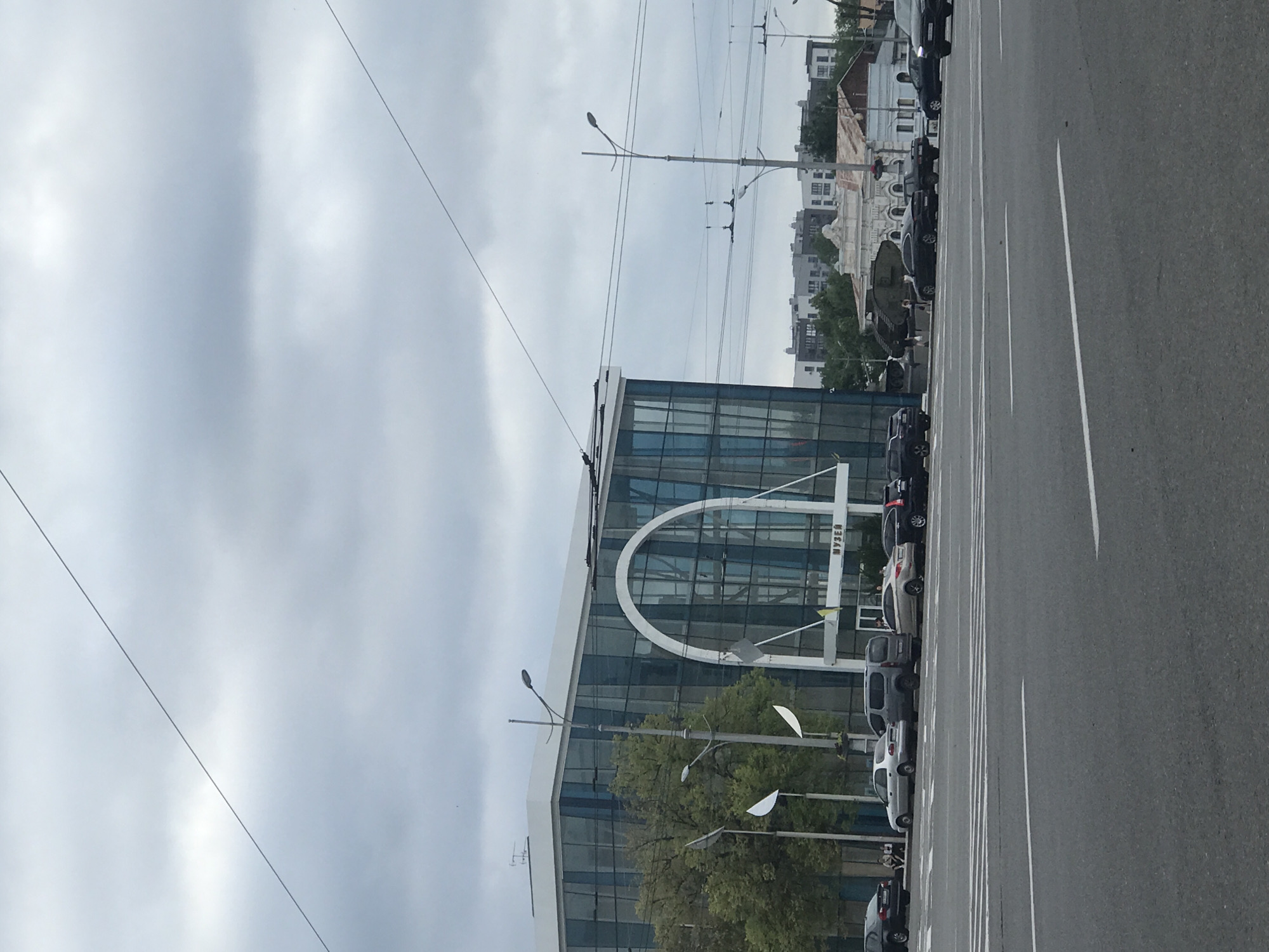This is a medium-distance, eye-level, street-level shot taken on a cloudy day, likely in Kharkiv, Ukraine. The focal point is a modern, multi-story building with a distinctive arched entrance and blue-tinted glass facade. Text above the entrance reads "МУЗЕЙ", indicating it is a museum. In front of the museum, several cars are parked along the street.
To the left of the museum, trees provide a natural element. A street lamp is positioned prominently in front of the building. Further down the street, various other buildings and structures can be seen, including what appears to be a military tank displayed outdoors behind a fence. The foreground of the image is dominated by a wide asphalt road with visible lane markings and a pedestrian crosswalk. Several cars are driving or parked on the road and adjacent to the museum. A few individuals are seated on a bench near the museum's entrance. Overhead, electrical wires stretch across the sky, suggesting an urban setting with public transportation infrastructure. The overall atmosphere is overcast, contributing to a somewhat muted color palette.
