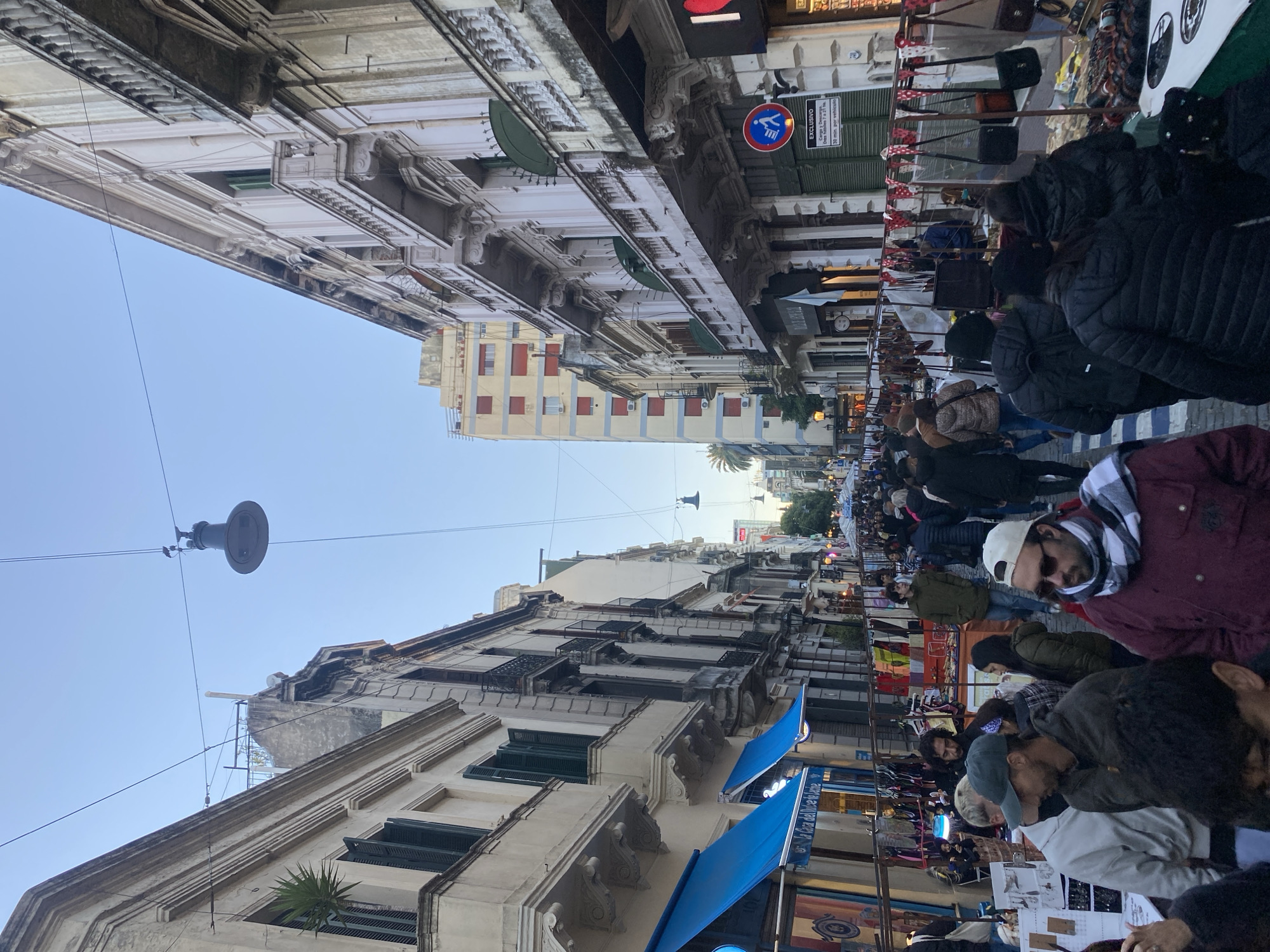 The image shows a bustling street scene in Buenos Aires, Argentina. The architecture is ornate, with old buildings lining both sides of the street. Blue awnings are visible in front of some of the businesses. A dense crowd of people is walking down the street, likely browsing the market stalls set up along the sides. These stalls display a variety of goods, including clothing, bags, and other items. The sky is blue, suggesting it is daytime. Overhead, streetlights are suspended by wires between the buildings. The image captures the lively atmosphere of a market day in Buenos Aires.