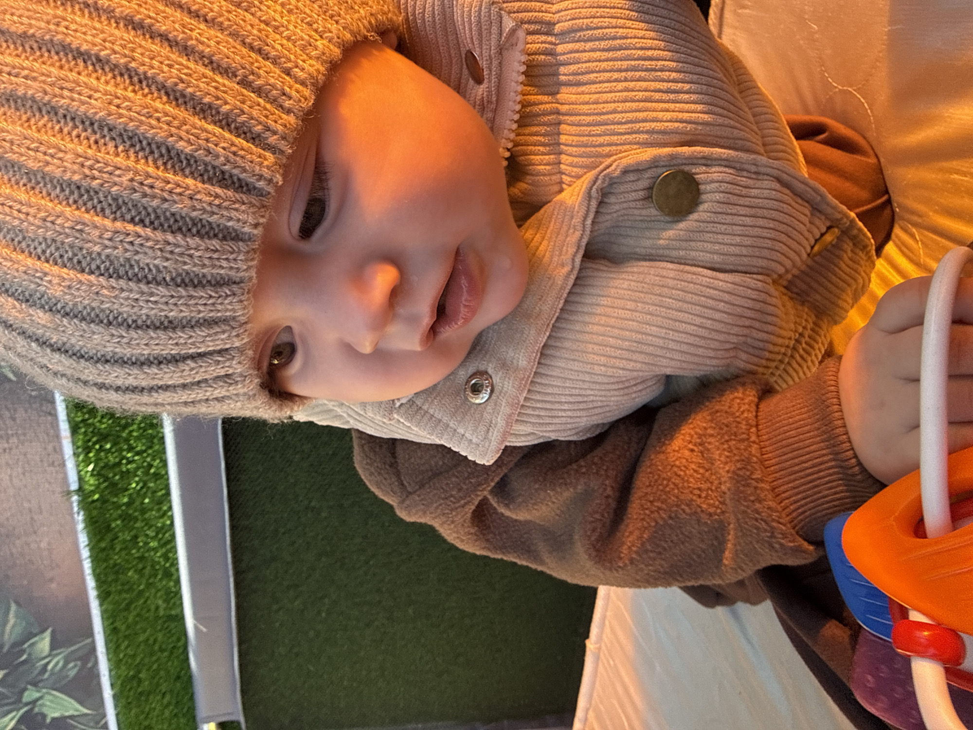A baby wearing a knit hat and a corduroy jacket is lying on a white surface. The baby is holding a colorful toy. The background includes a patch of artificial turf. The location is unknown, and there is no verifiable information to indicate it is Кипчак, Russia.