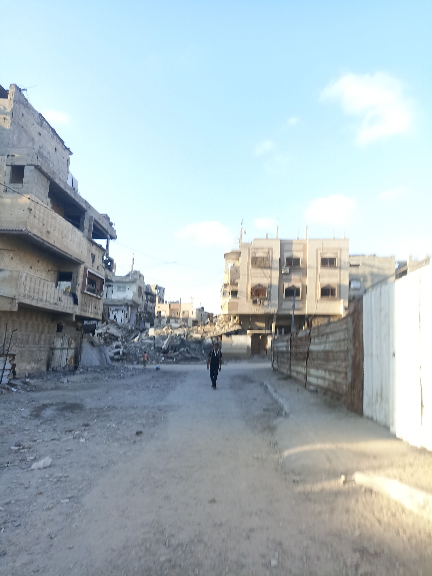 The image depicts a street scene in a city with evident signs of destruction. On the left, a multi-story building displays significant damage, with exposed concrete, missing walls, and bullet holes. Debris, including rubble and construction materials, is piled high in the middle ground, indicating the aftermath of an event such as an explosion or bombardment. Further down the street, more damaged buildings are visible, some appearing partially collapsed.
A person, dressed in dark clothing, walks away from the camera down the center of the unpaved road. To the right, a fence or wall constructed from corrugated metal panels runs along the side of the street, with a white structure behind it. The sky is clear and blue with a few scattered clouds. The overall impression is one of urban devastation and desolation.