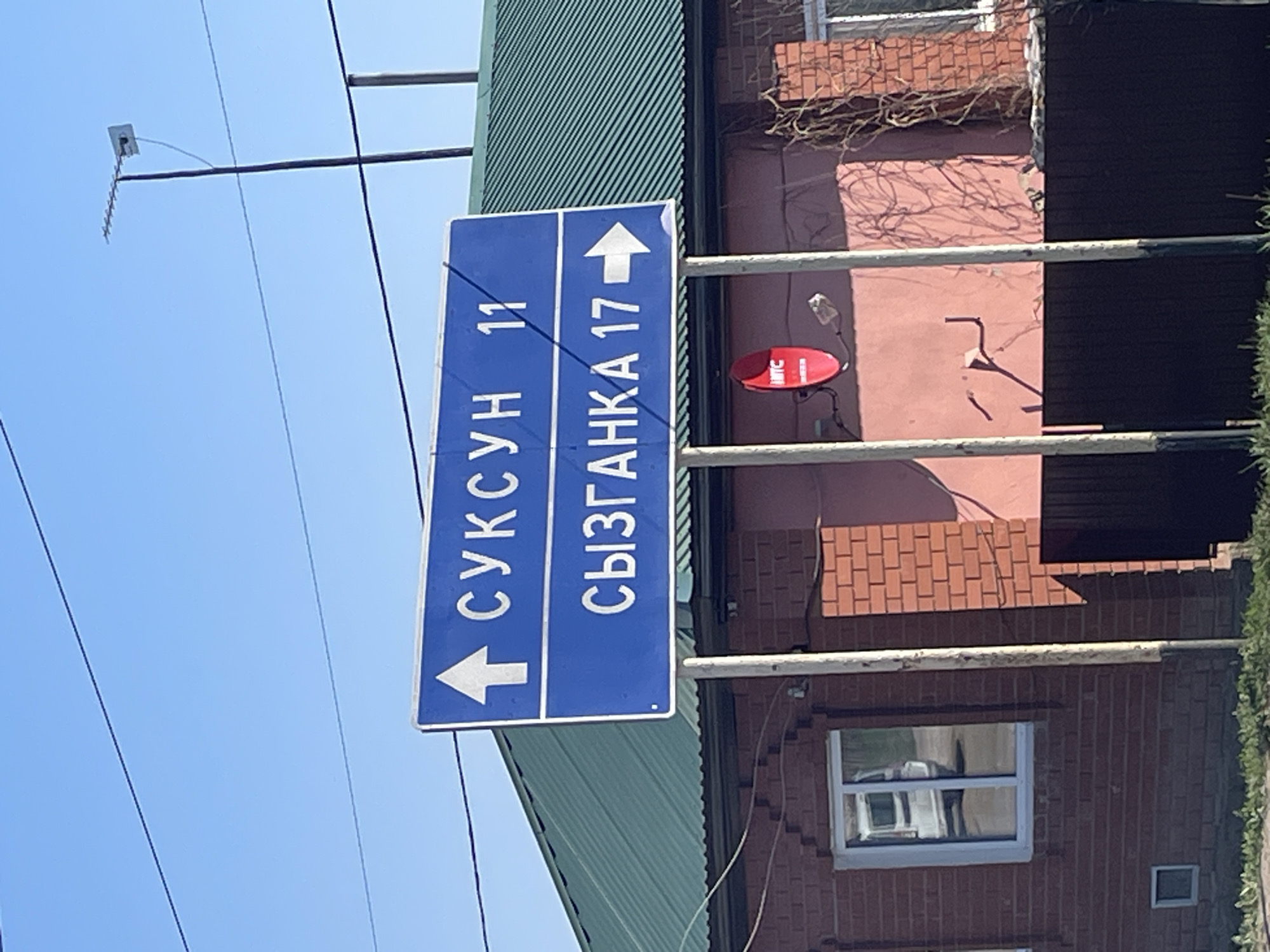 A medium shot shows a blue street sign in Поедуги, Russia. The sign is vertically oriented with white lettering indicating two destinations and distances. To the left with an arrow pointing left, it says "СУКСУН 11". To the right with an arrow pointing right, it says "СЫЗГАНКА 17". The sign is affixed to a metal pole.
Behind the sign, there is a building with a green corrugated metal roof and a brick facade. A red satellite dish is visible on the wall. A window is present on the brick wall of the building.
In the upper left of the frame, there are utility lines and an antenna against a clear blue sky. The image appears to be taken during the daytime.