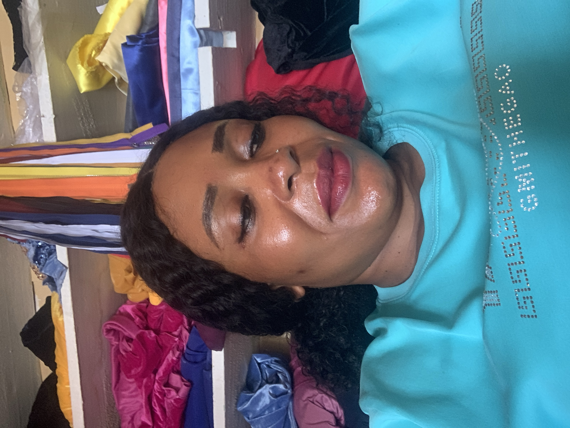 A woman with dark curly hair and a nose piercing is lying down wearing a light blue sweatshirt with studded lettering. Behind her is a rack of colorful fabrics, including yellow, pink, blue, red and black. The location appears to be Sabon Tasha, Nigeria, based on visual cues.