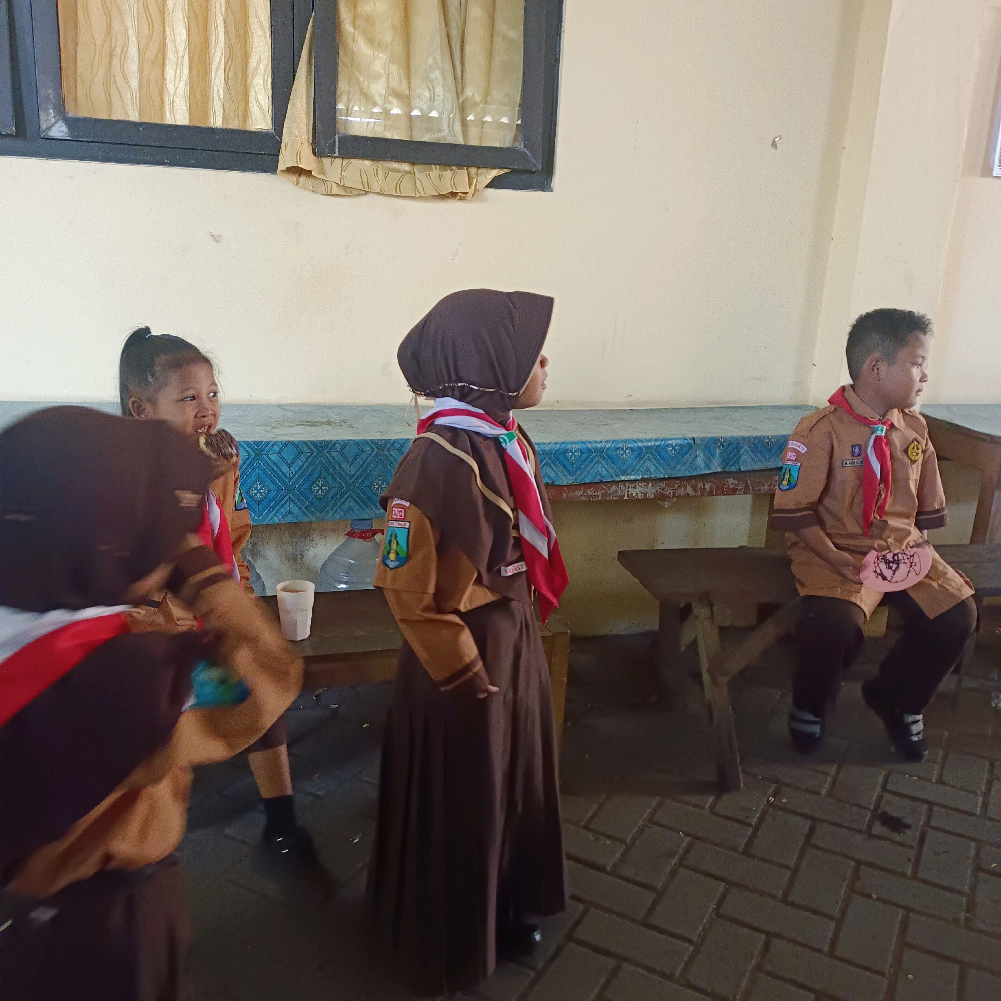The image depicts a group of young individuals dressed in scout uniforms, likely on the grounds of an educational institution in Surabaya, Indonesia. The setting appears to be outdoors, with a pale, textured wall and a window with beige curtains in the background. The ground is paved with interlocking bricks.
In the foreground, on the left, a person in a brown scout uniform with a red and white scarf is partially visible, with their back to the camera. They are wearing a dark headscarf. To their right, a girl with her hair in a ponytail is seated, also in uniform, eating what appears to be a donut. She is looking to her right.
In the middle ground, a girl wearing a dark headscarf and a brown scout uniform with a red and white scarf is standing and looking to her right. She has a badge on her left sleeve. To her right, a boy in a similar brown scout uniform with a red scarf is seated on a wooden bench. He is looking to his right and holding a pink paper object. He has a badge on his left sleeve as well.
Various objects are present, including a white cup, a plastic water bottle, and wooden benches. The overall atmosphere suggests a casual gathering or break during a scouting activity. The uniform colors and badges suggest a specific scouting organization, possibly associated with a school in Surabaya.
