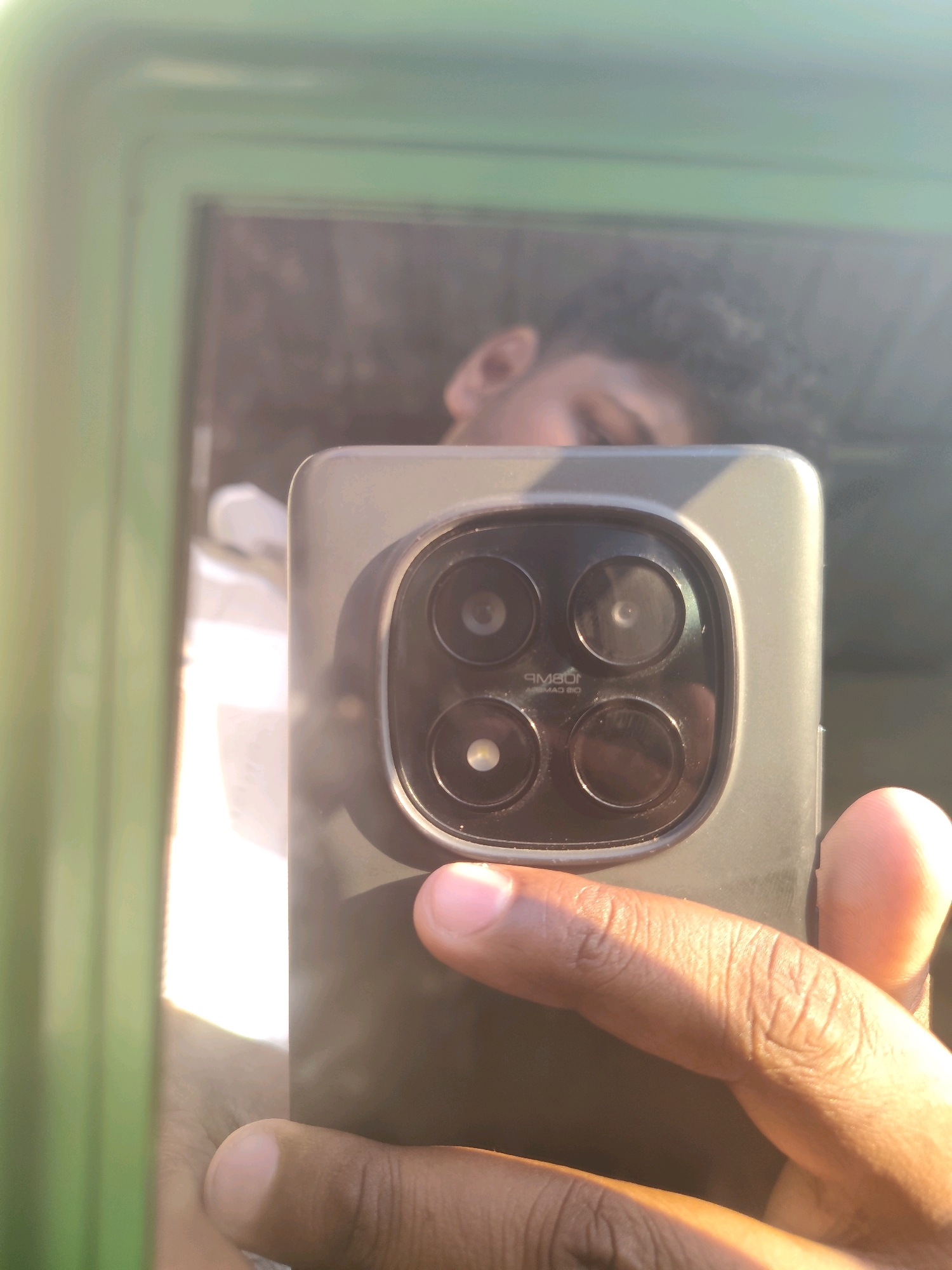 A person's hand is holding a smartphone in a selfie, capturing a reflection of their face in a mirror. The person appears to be young with dark, curly hair and is wearing a white shirt. The smartphone has a prominent camera module with four lenses arranged in a circular pattern. Text is visible on the camera module, reading "108MP CAMERA". The background shows a bright, outdoor environment with a green, textured surface on the left, likely part of a structure or wall. The scene suggests a casual moment, possibly in an urban setting. The reflection in the mirror shows the person's upper body and face, with their eyes looking towards the camera. The lighting indicates it is daytime, with sunlight casting shadows and creating highlights on the phone and hand. The reflection also shows a faint outline of what might be another person or object behind the subject, further suggesting an outdoor or semi-public space within the city of Belagavi, India.