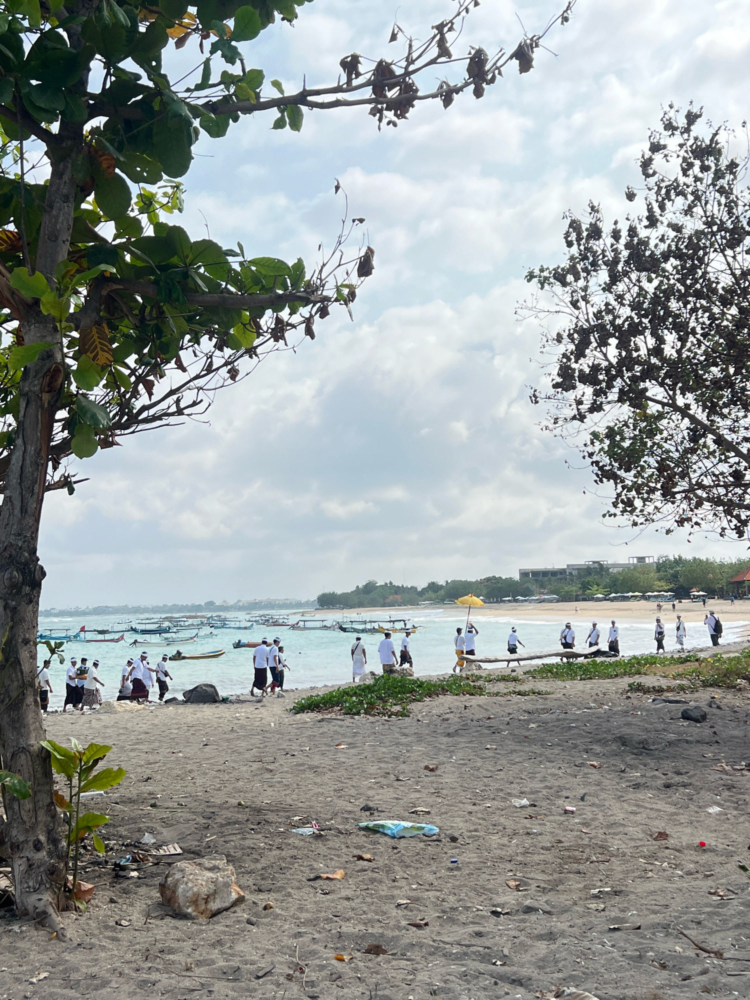 In Kuta, Indonesia, a group of people dressed in traditional Balinese attire, consisting of white shirts and sarongs, are walking along the beach, with some carrying offerings. They are accompanied by a person holding a yellow umbrella. The beach has dark sand with scattered debris, including plastic bottles and wrappers. In the background, a line of traditional Balinese fishing boats with outriggers are moored in the turquoise ocean. Beyond the boats, a coastline with buildings and lush green trees is visible under a cloudy sky. The scene is framed by the branches of a tree in the foreground on the left and a bare tree on the right.