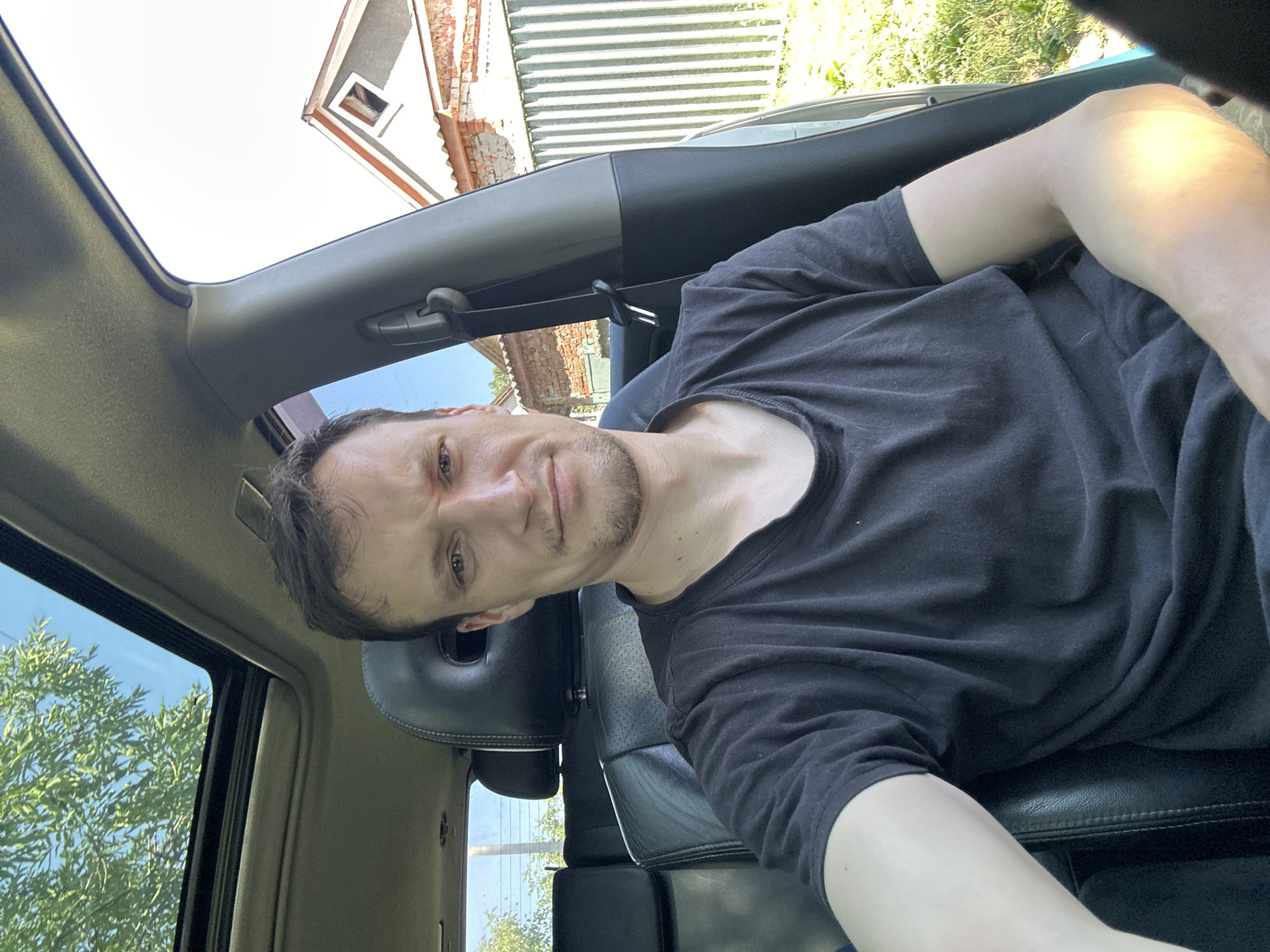 A man is seated inside a vehicle. He is wearing a black t-shirt and has short brown hair and stubble. The man is looking directly at the camera with a neutral expression. Sunlight is coming in through the sunroof of the car, casting a bright light on his right arm. The interior of the car is visible, including black leather seats and the car's dashboard. Outside the car, a residential building with a red-tiled roof and a green metal fence is visible. Trees and grass are also present in the background, suggesting an outdoor, possibly rural or suburban, environment. The image was taken in the city of Makiv, Ukraine.
