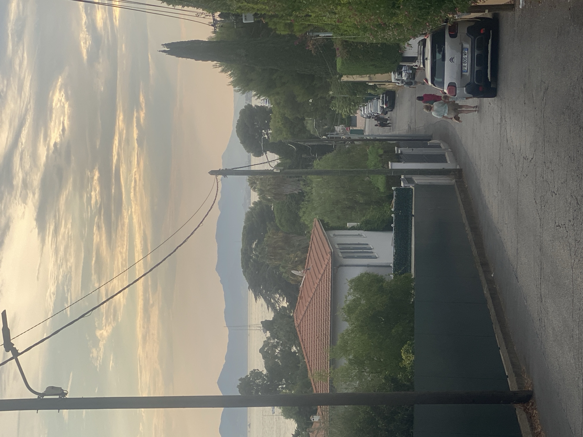The image depicts a street scene in Antibes, France, looking downhill towards the sea and distant mountains. A tall utility pole with a streetlamp and wires dominates the left foreground. The street itself is paved and slopes downward, with parked cars lining both sides. On the left, a white house with a red tile roof is visible behind a dark green fence. Further down the street, several people are walking, including two women in the foreground. The right side of the street is lined with dense green foliage and trees. The sky above is partly cloudy with a soft, hazy light, suggesting either early morning or late afternoon. In the distance, sailboats can be seen on the water, with a mountain range forming the horizon. The license plate of the silver car in the foreground reads "FR 806 WP."