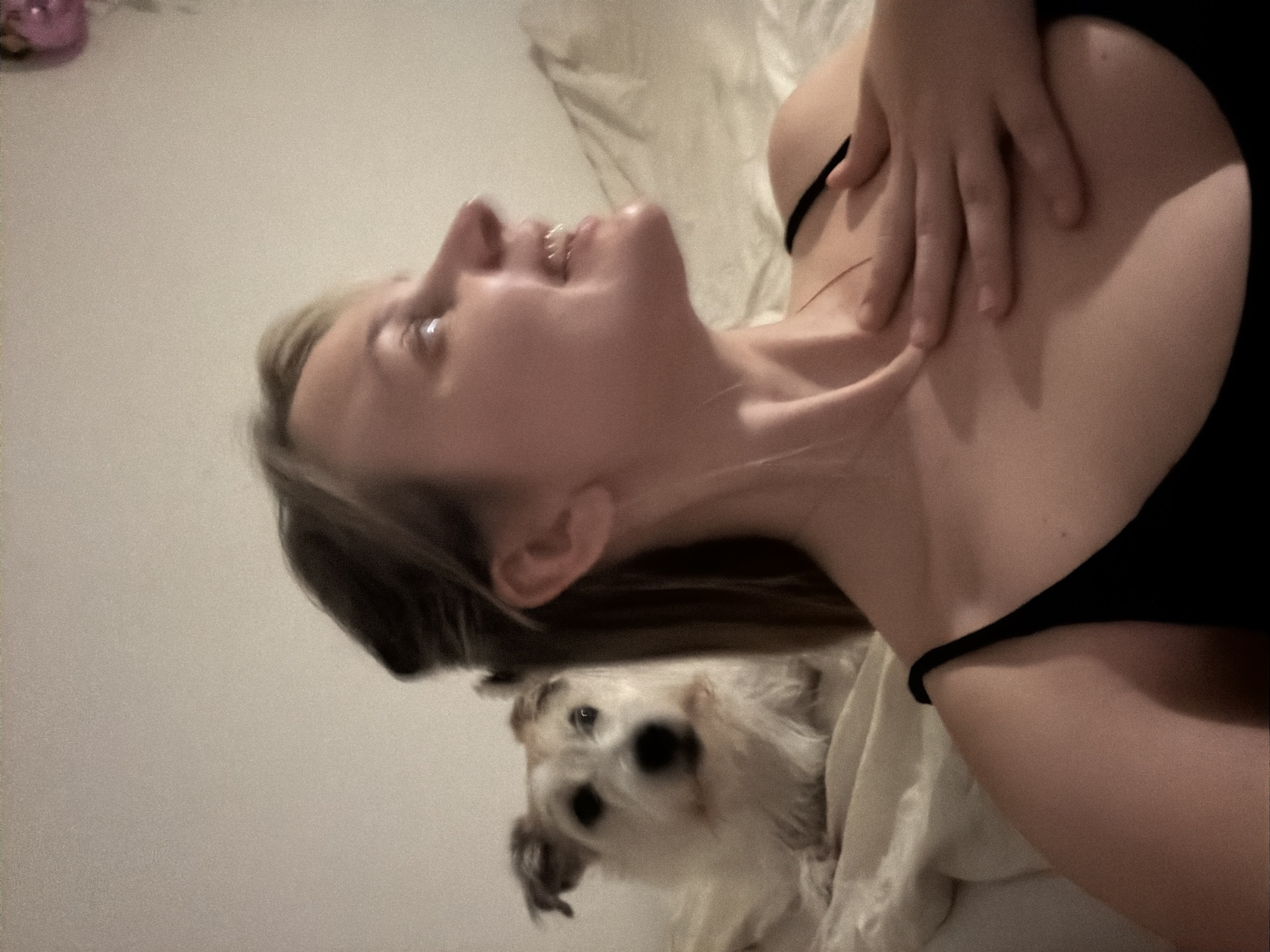 A woman with blonde hair is lying on a bed, looking upward and smiling. She is wearing a black tank top. Her hand is on her chest. A small, white dog is at the foot of the bed, looking toward the camera. The background is a plain wall. There is no indication this is in Tel-Aviv, Israel.