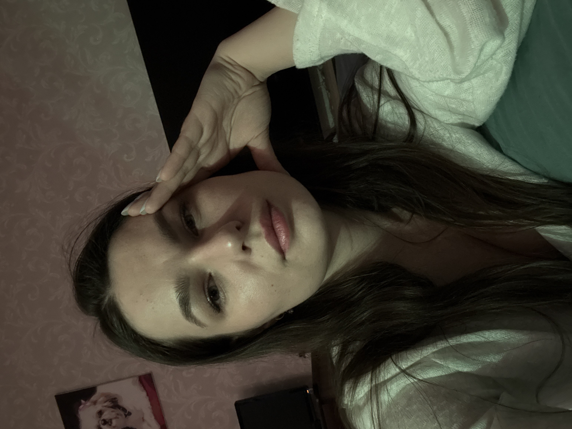 The image features a young woman, positioned at an angle, looking directly towards the viewer. Her long, dark hair frames her face and spreads across her shoulders and chest. Her right hand is placed on her forehead or temple, with light-colored, possibly pastel blue or green, nail polish visible on her fingers. She is wearing a light-colored, possibly white or off-white, textured top or blanket.
The woman's expression is neutral to slightly pensive, with a subtle pout to her lips. The lighting is warm and artificial, casting shadows and suggesting an indoor setting, likely in the evening or at night.
The background consists of a wall covered in light pink or beige wallpaper featuring a subtle, intricate floral or damask pattern. To the upper right, a dark area transitions into a teal or green fabric, possibly a blanket or clothing. Below the woman's face, a small framed picture is visible on the patterned wall, depicting a fluffy white animal, possibly a dog or cat, with some reddish hues. A dark, rectangular object, potentially a phone or remote control, is partially visible near the bottom edge of the frame.
There is no visible text in the image. The location context provided is Chaikovsky, Russia.