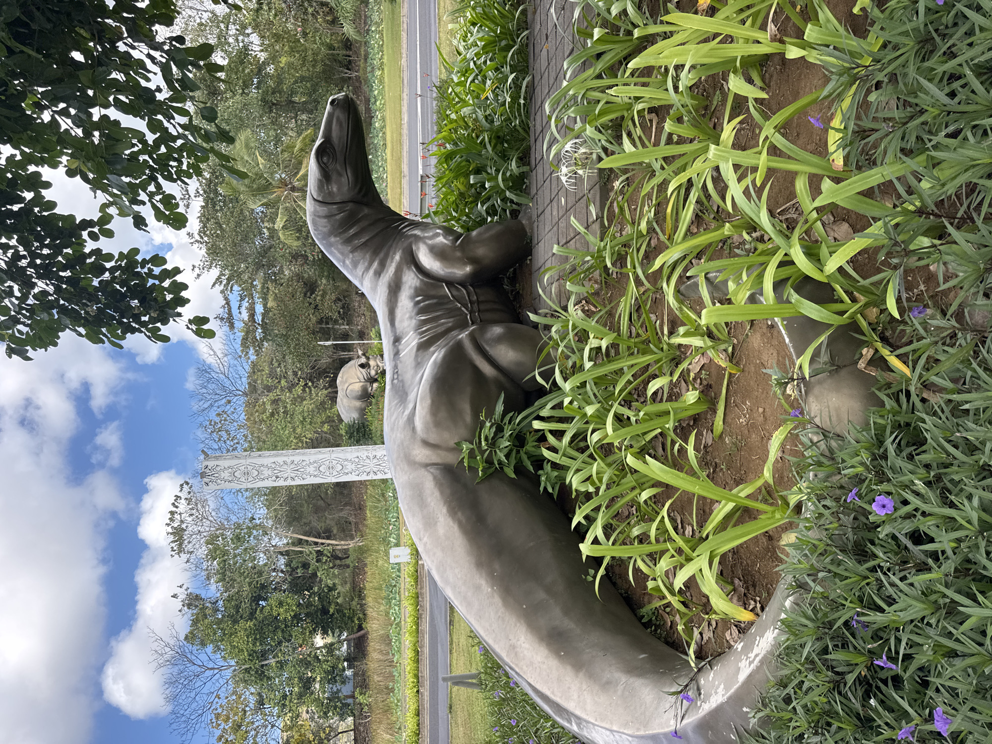 This image depicts a large, metallic sculpture of a monitor lizard in an outdoor setting. The sculpture is dark gray and detailed, showing its scaly skin and powerful limbs. It is positioned in a garden bed surrounded by lush green foliage and small purple flowers. In the background, a road with some passing traffic is visible, along with trees and a cloudy blue sky. Further in the background, another animal sculpture, possibly a rhino, can be seen near a tall, decorative pillar. The overall scene suggests a public space, possibly a park or an entrance to a tourist attraction in Kuta Selatan, Indonesia.