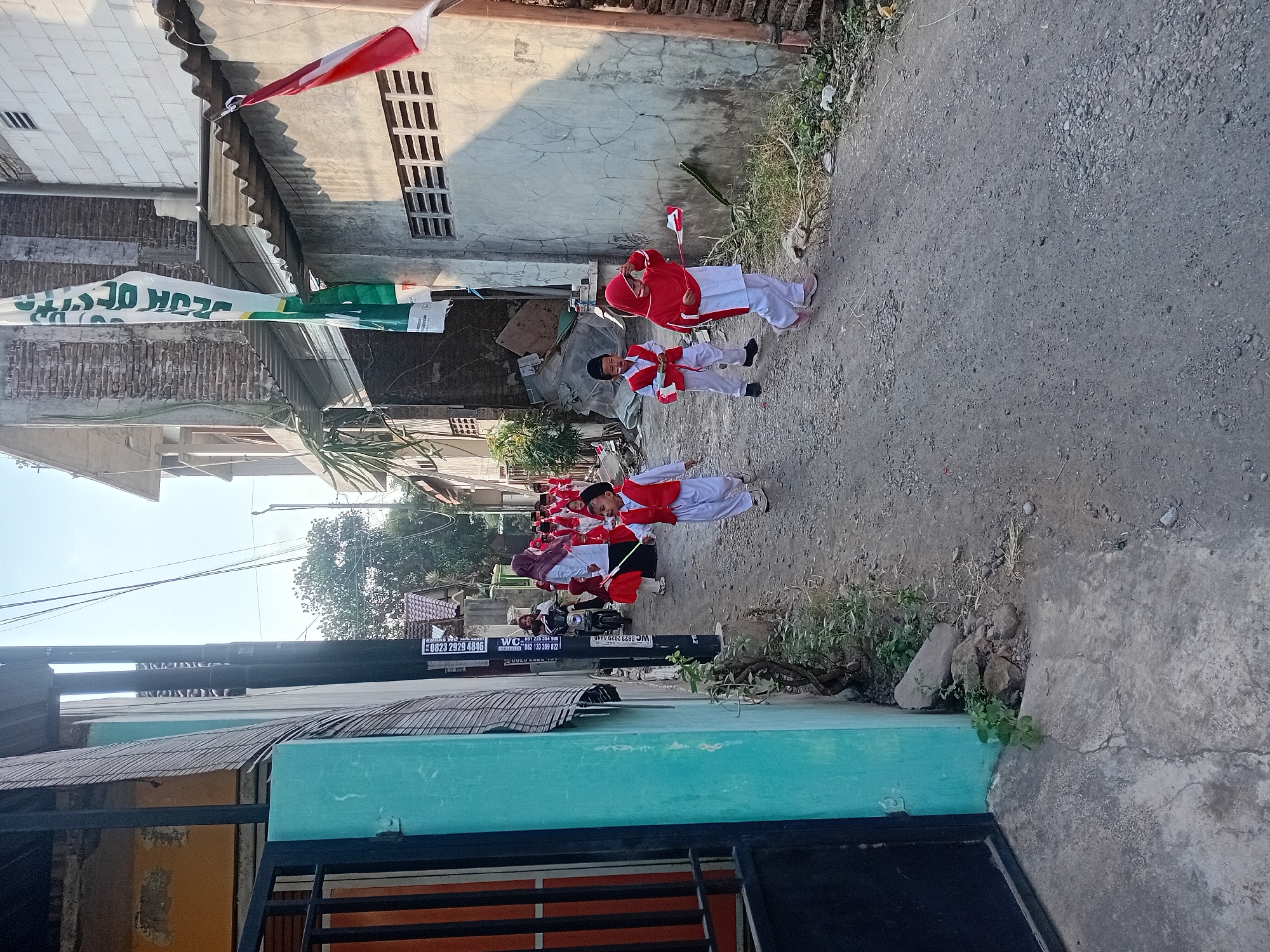 In Besito, Indonesia, a group of young children, dressed in red and white uniforms and hats, are participating in a parade on a narrow, unpaved street. They are carrying small Indonesian flags. A woman on a motorcycle is visible in the background, and several buildings line the street, some of which are adorned with flags and banners. The scene appears to be a community event, possibly a celebration or holiday, under a clear sky.