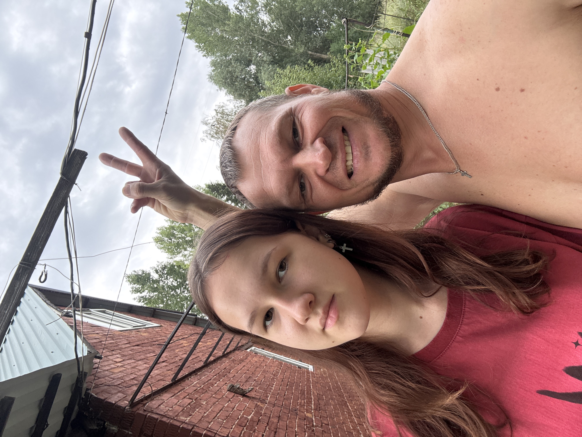 A young female with long brown hair and a red t-shirt poses for a selfie with a shirtless man. The man is smiling and making a rock-on hand gesture. Behind them is a red brick building and trees. The image appears to be taken outdoors, possibly in a rural or suburban setting. There is no indication of the city of Oskemen, Kazakhstan.
