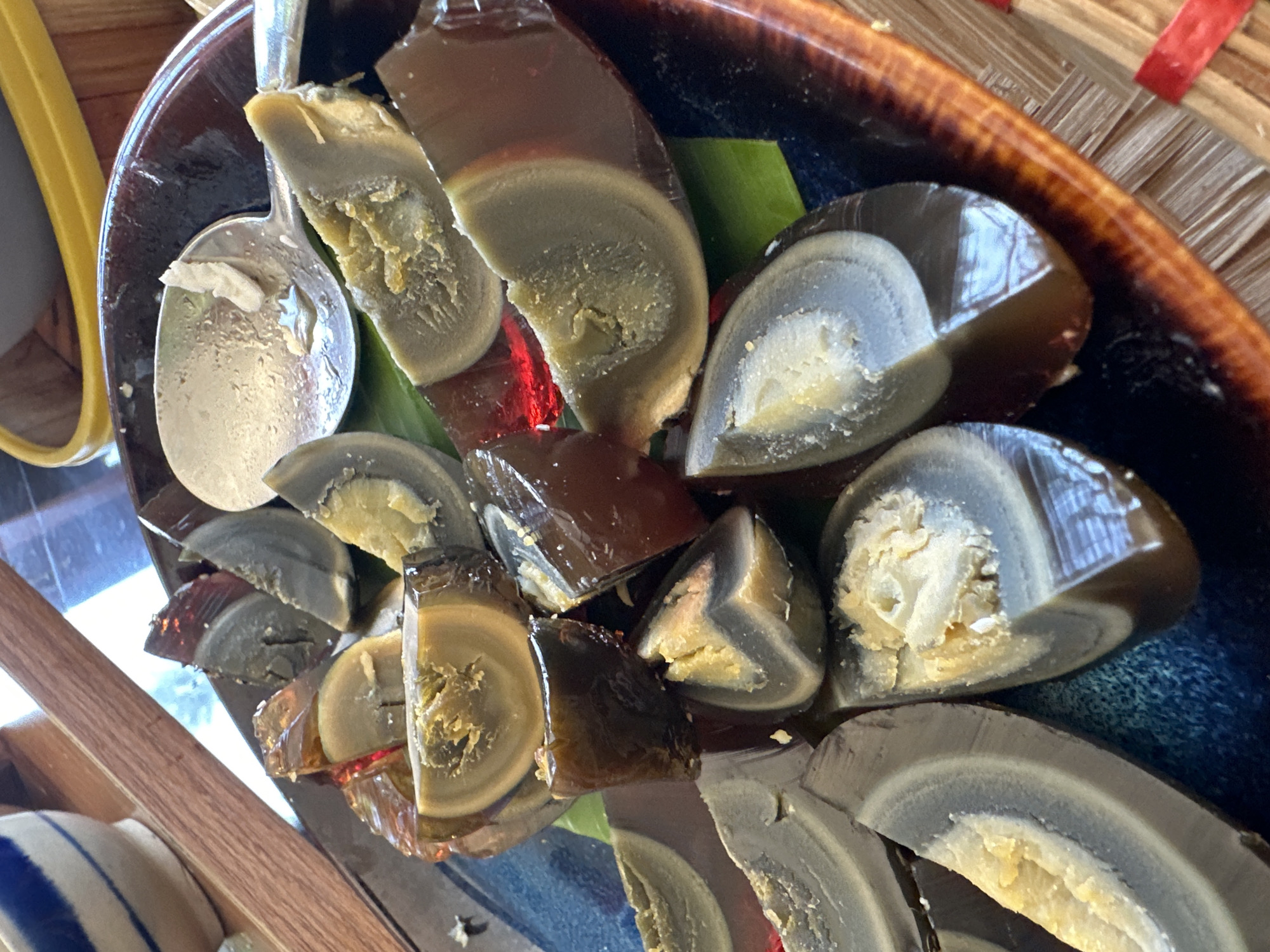 The image displays a close-up, high-angle view of a serving of prepared century eggs on a dark, round plate. Approximately two dozen pieces of cut century eggs are visible. Each piece is typically a wedge or half-section, revealing the internal structure. The albumen (egg white) is dark brown, translucent, and gelatinous, with some segments exhibiting a reddish tint when light passes through. The yolk varies in appearance; some are dark greenish-grey and firm, while others are a granular, pale yellow-brown, sometimes displaying a darker ring around the core. Concentric layers are discernible within the albumen of several egg pieces. Some egg sections are resting on fragments of a green leaf, possibly a banana leaf, on the plate.
The eggs are served on a dark, possibly ceramic or glazed clay, round plate with a distinct reddish-brown, textured rim. A silver-colored metal spoon is partially visible in the upper right, containing a small amount of light-colored, granular residue.
In the background, a white ceramic bowl with vertical blue stripes is partially visible in the upper left. A wooden surface or plank runs diagonally across the upper left and center of the frame. The yellow rim of another dish or container is present in the upper right. In the lower right, a woven surface, appearing to be a mat made of light-colored natural fibers with a red band, is partially visible. The scene is illuminated by ambient light, causing reflections on the translucent egg albumen and the spoon. No individuals, direct actions, or interactions are depicted.
The specific geographic location in Nha Trang Ward, Vietnam, cannot be determined from the visual information provided in the image.