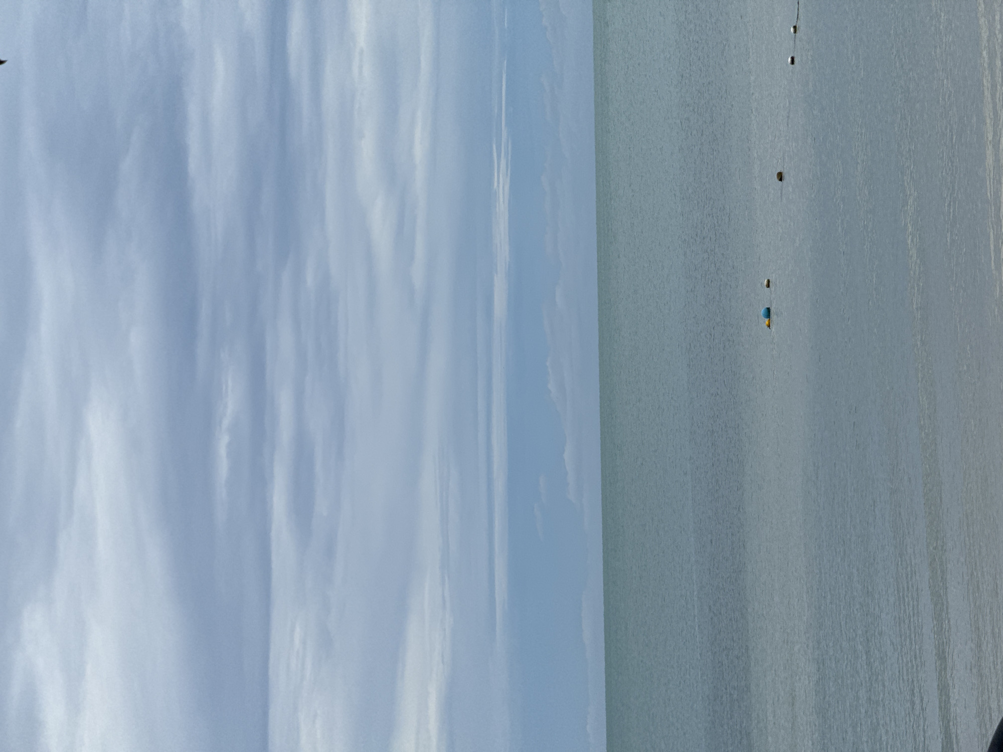 A wide shot shows a body of water with a cloudy sky above. The water is calm and a light, muted blue-green. Several buoys are visible in the water, including a yellow and blue one in the mid-ground, and a series of smaller, spherical buoys further out. The sky is filled with layers of grey and white clouds, with some lighter blue patches showing through. The horizon line is clearly defined, separating the water from the sky. There are no people or buildings visible in the frame. The overall mood is serene and slightly overcast.