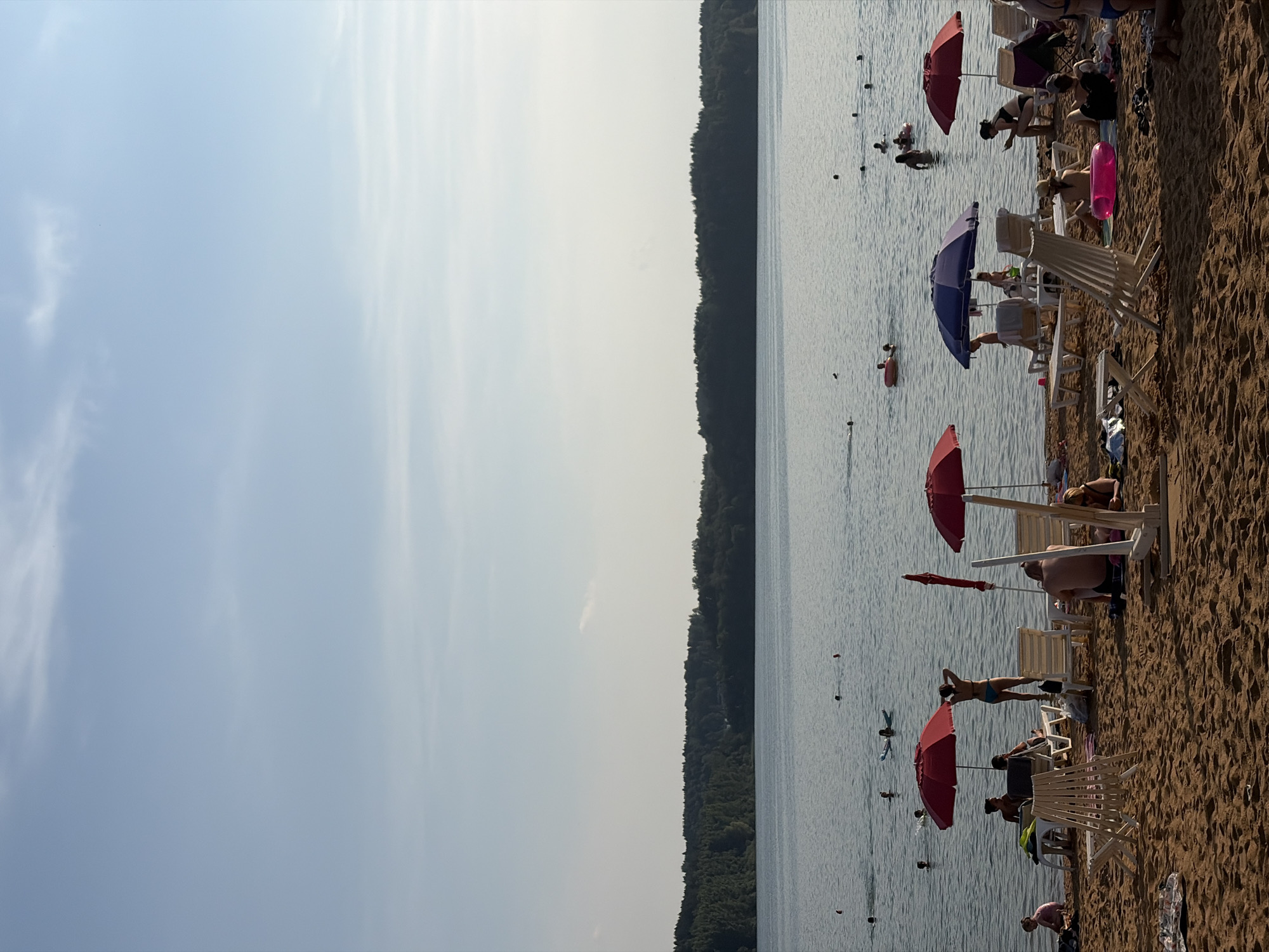A wide shot captures a beach scene on a body of water, possibly a lake, with a treeline in the background. The sky is a clear, pale blue with faint wisps of clouds. The water is calm, with several people swimming or wading. On the sandy beach, various individuals are sunbathing, relaxing on lounge chairs, or walking. Red and blue umbrellas provide shade for some beachgoers. The overall impression is a typical summer day at a recreational area. This location is identified as being in the Ozyaritska-Slabadski rural council, Belarus.