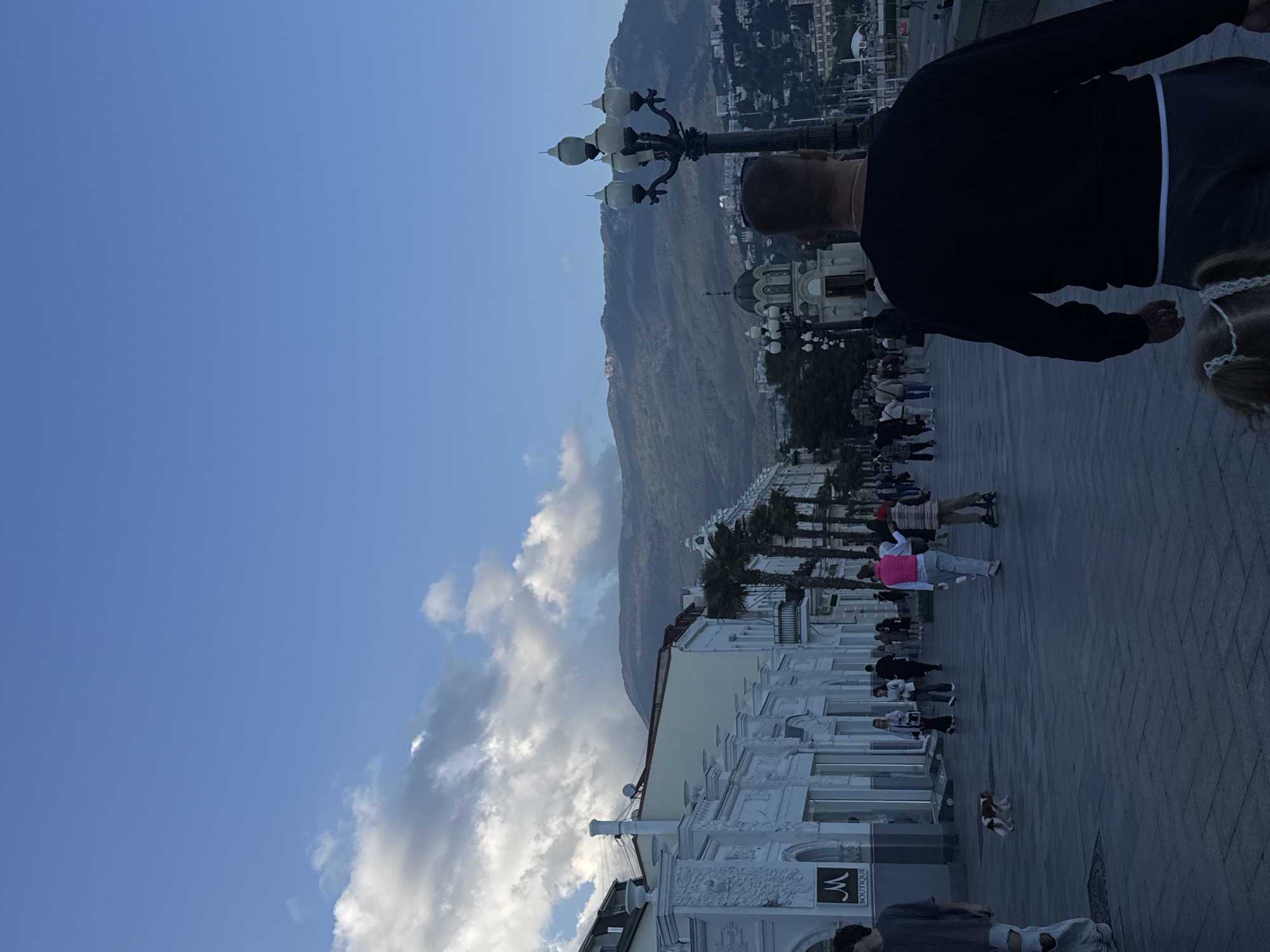 The image shows a street scene in the city of Yalta, Ukraine. Buildings with white facades and intricate architectural details line the street. A mountain range forms the backdrop. The sky is blue with scattered clouds. People are walking along the paved street. There is an ornate lamppost. A dog is visible on the street. A man in a black jacket and grey pants is partially obstructing the view.