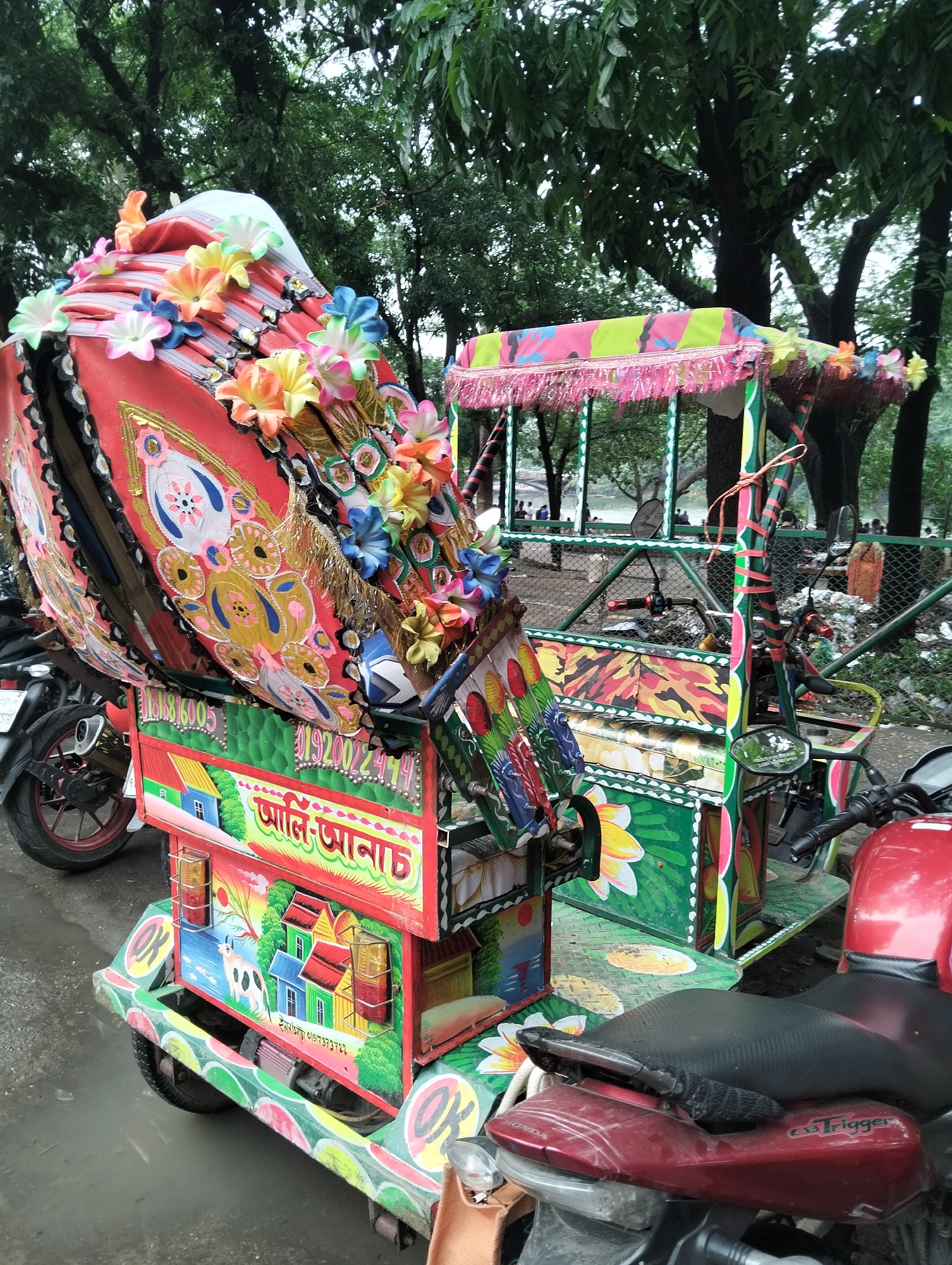 A colorfully decorated auto-rickshaw or battery-powered rickshaw (easy bike) is parked on an asphalt surface in what appears to be a park or outdoor recreation area in Dhaka, Bangladesh. The rickshaw features vibrant and intricate hand-painted designs, including floral motifs and depictions of rural scenes with houses and animals. The rickshaw's canopy is adorned with artificial flowers. Other parked motorbikes surround the rickshaw. In the background, trees and a fenced-off area with people and potential waste accumulation can be seen. The sky appears overcast.