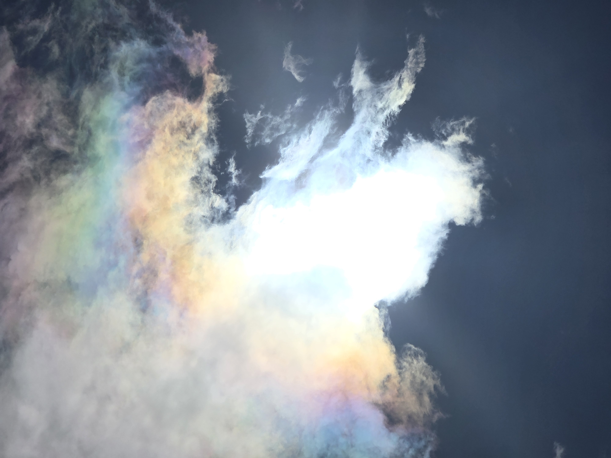 A high-angle, eye-level shot captures a dramatic display of iridescence in a dark sky, likely over the city of Ермолаево, Russia. The sky is a deep, almost black blue, with wispy, white clouds scattered throughout. These clouds are illuminated by a very bright, white light source, likely the sun, which is obscured by a large, dense cloud formation. This bright light is refracting through the ice crystals within the surrounding clouds, creating a vibrant, iridescent halo of colors that includes shades of pink, yellow, green, blue, and purple. The iridescence is most prominent in the upper left quadrant of the image, bleeding into the white clouds and then gradually fading into the darker sky. The clouds themselves appear thin and ethereal, with some areas being dense and opaque, while others are translucent, allowing the colors to shine through. The overall impression is one of a natural phenomenon, a beautiful and fleeting display of light and color in the atmosphere. There are no people or man-made objects visible in the image.