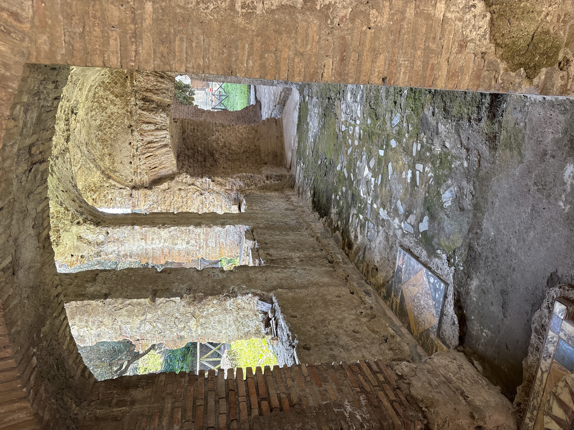 The image shows an interior view within Hadrian's Villa in Tivoli, Italy. The space is constructed of aged brick and stone, exhibiting significant decay. Natural light penetrates through openings in the structure, illuminating the interior. There is exposed masonry and visible signs of aging such as moss growth and crumbling material. A fragment of decorative tilework can be seen set within one of the wall sections, indicating the original presence of more elaborate design. Views to the exterior through the dilapidated structure reveal foliage and scaffolding. No people are visible.