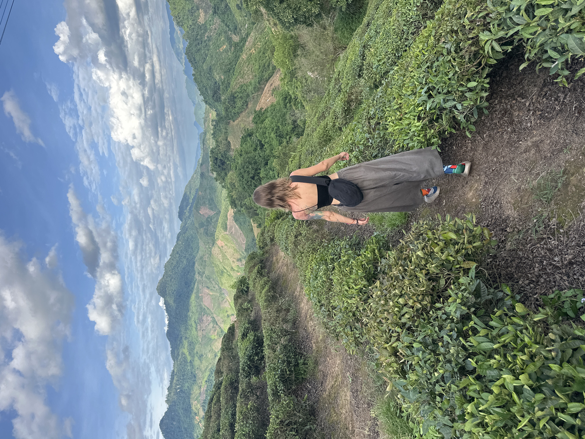 The image depicts a woman walking on a dirt path through lush green tea fields on a hillside. She is viewed from behind, wearing a black crop top, a long olive green skirt, and colorful striped socks with athletic shoes. A small black crossbody bag is slung over her shoulder. The tea fields are neatly cultivated, forming terraced rows that cascade down the mountainous terrain. In the background, a vast panorama of rolling green hills and mountains stretches towards a sky filled with white, fluffy clouds under a bright blue sky. The location is identified as To Mua, Vietnam, a region known for its scenic tea plantations. The overall scene suggests a serene and picturesque natural landscape.