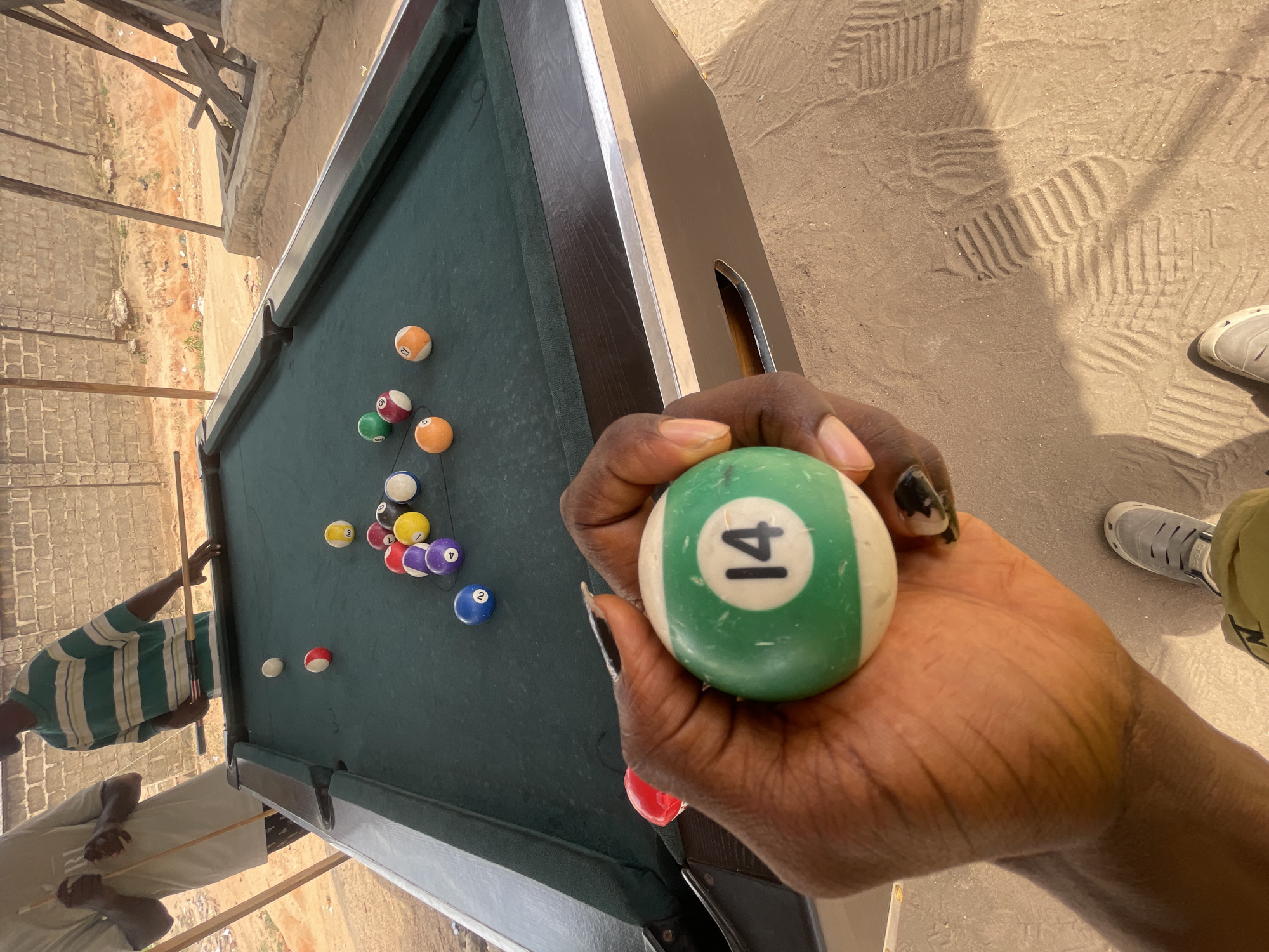 The image shows a hand holding a green and white billiard ball with the number 14 on it. The billiard ball appears worn. The hand is dark-skinned and the fingernails have dark polish. Behind the hand is a billiard table with several other billiard balls scattered on the green felt surface. A person wearing a green and white striped shirt is partially visible near the edge of the table. The location appears to be outdoors. There are hints of urban development near the table in what appears to be the city of Lokoja, Nigeria. A foot wearing a light-colored athletic shoe is partially visible near the bottom right corner of the image.