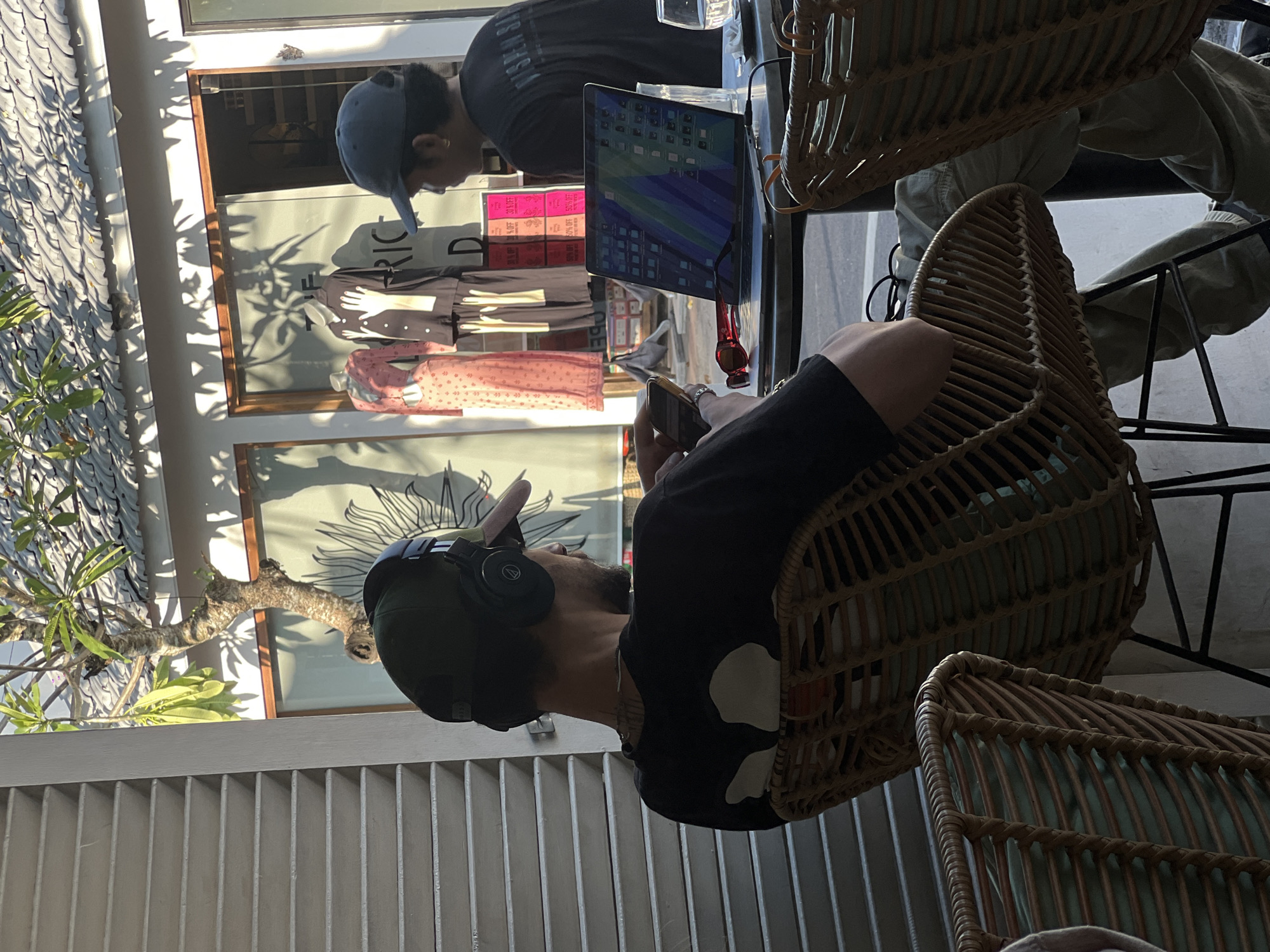 The photograph captures a scene at a café in what appears to be North Kuta, Indonesia. The perspective is from within the establishment, looking towards an outdoor area.
Inside, two individuals are seated in wicker chairs. One person wears a dark t-shirt with what appears to be white skull shapes, and has headphones around his neck. He is looking down at a mobile phone in his hands.
A second person is sitting at the same table, wearing a gray shirt and a baseball cap. A laptop is positioned on the table in front of them.
Through a glass storefront, various mannequins wearing clothing are visible. A sidewalk with some greenery is also present. Light conditions indicate it is daytime. The overall atmosphere seems relaxed and casual.