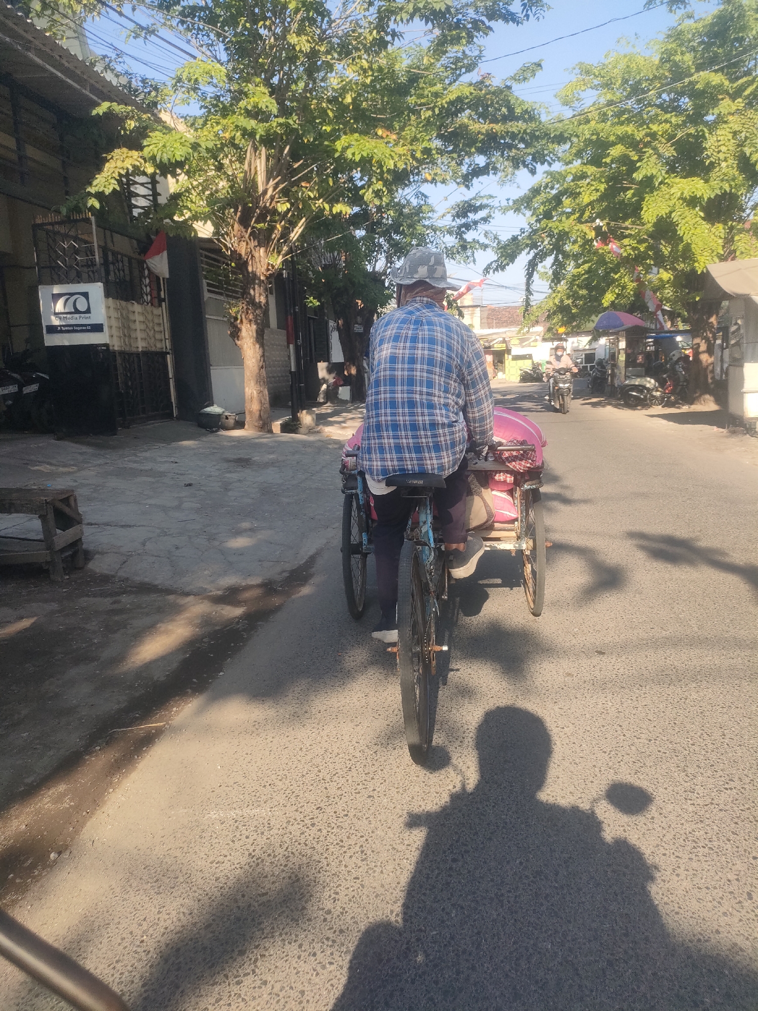 The image captures a street scene in Surabaya, Indonesia, bathed in the warm light of a clear day. In the foreground, a person is riding a bicycle, pulling a cart. The rider is wearing a blue and white plaid shirt and a patterned hat, seen from behind. The cart is partially obscured, but pink fabric is visible within it. The bicycle has a metal frame and spoked wheels. The road surface is asphalt, with shadows cast by trees and the cyclist's shadow prominent on the right side of the frame.
To the left of the road, a sidewalk is visible, with a wooden bench or table. Behind the sidewalk, there are buildings. One building prominently displays a sign for "CV Media Print," with an address below it: "Jl. Tumam Segaran 63." Trees with lush green leaves line the street, some with small red and white flags strung between them.
In the background, other vehicles are present on the road, including motorcycles. One person on a motorcycle is visible further down the street, wearing a face mask and a helmet. The environment appears to be an urban or suburban area with a mix of commercial buildings, residential structures, and abundant greenery. The overall atmosphere is one of everyday life in a bustling Indonesian city.