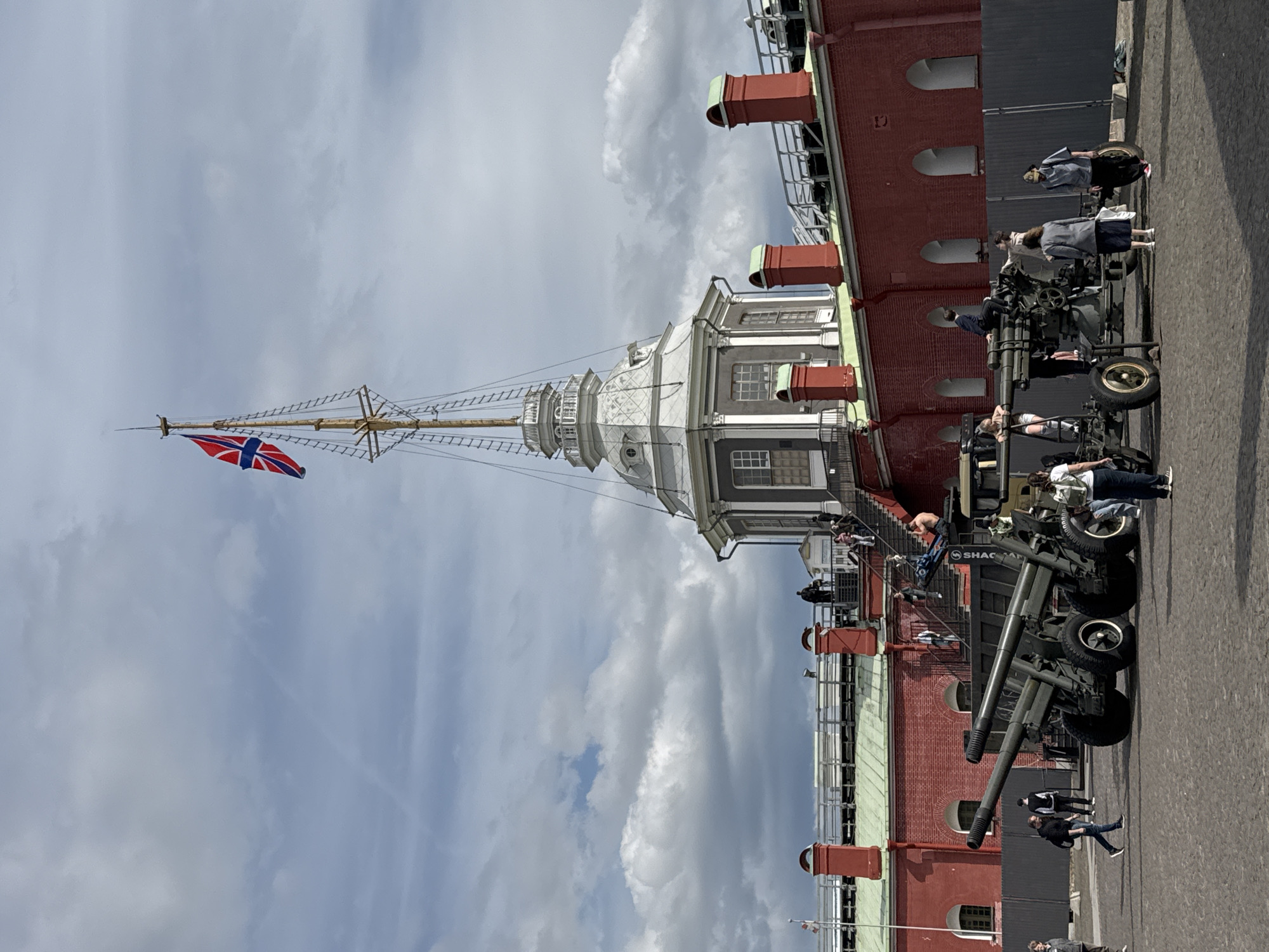 The image depicts an outdoor scene at the Peter and Paul Fortress in Saint Petersburg, Russia, under an overcast sky. A prominent multi-story red-brick building with arched window openings and green-trimmed eaves forms the backdrop. Central to the composition is an ornate white tower with a cupola, from which a tall wooden mast extends upwards. A blue flag with a red cross bordered in white, resembling the Union Jack, flies from this mast.
In the foreground and midground, several dark-green artillery cannons on wheels are positioned on a paved area, serving as exhibits. Various people are seen interacting with these historical weapons, some standing and observing, while others are seated or leaning on them. Additional visitors are visible on staircases that lead to higher sections of the brick building. The ground is paved, suggesting a public square or pathway within the fortress complex. The word "SHAC" is partially visible on the side of one of the cannons. The scene captures general tourist activity during what appears to be a cloudy daytime.