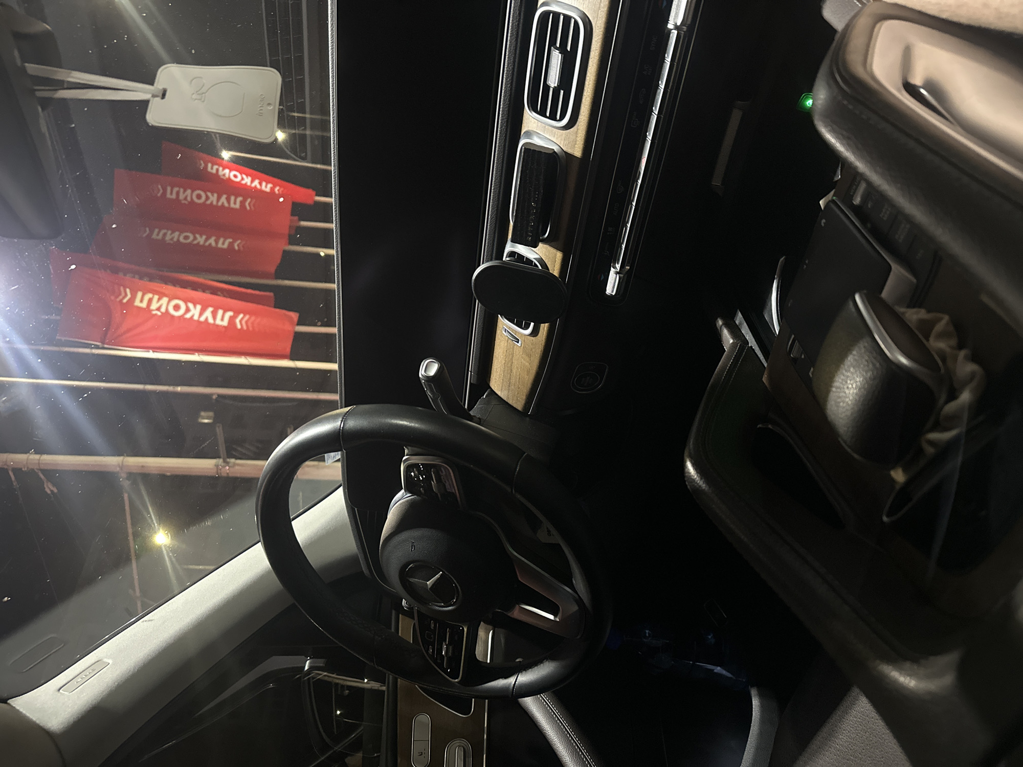 The image shows the interior of a modern car, likely a Mercedes-Benz, at night. The dashboard is visible, featuring a prominent steering wheel with the Mercedes-Benz logo. Wood-grain trim accents the dashboard and center console. A phone mount is attached to the air vent. In the background, through the windshield, several red flags with white Cyrillic lettering are visible, along with a white air freshener hanging from the rearview mirror. The flags appear to bear the logo "ЛУКОЙЛ," a Russian energy company. The scene is dimly lit, with some light reflecting off the windshield. Based on the presence of "ЛУКОЙЛ" flags, this scene could be located near a Lukoil gas station or an event sponsored by the company in Ruse, Bulgaria. No people are visible in the image.