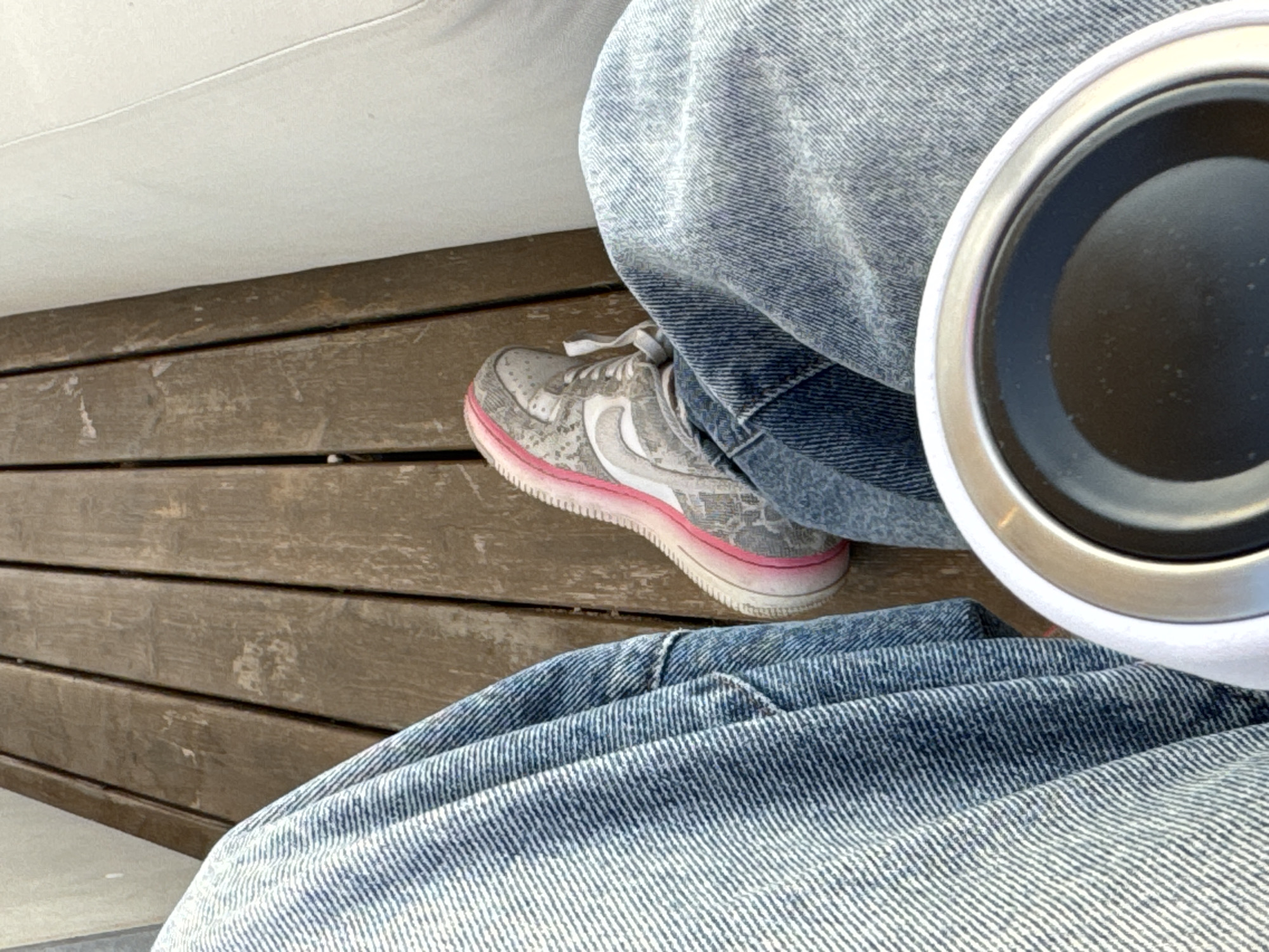 A person wearing blue denim pants and grey sneakers with a pink stripe on the sole is sitting on a wooden surface. A white cup or container with a black interior is visible in the foreground. The location is Мысовское сельское поселение, Ukraine.