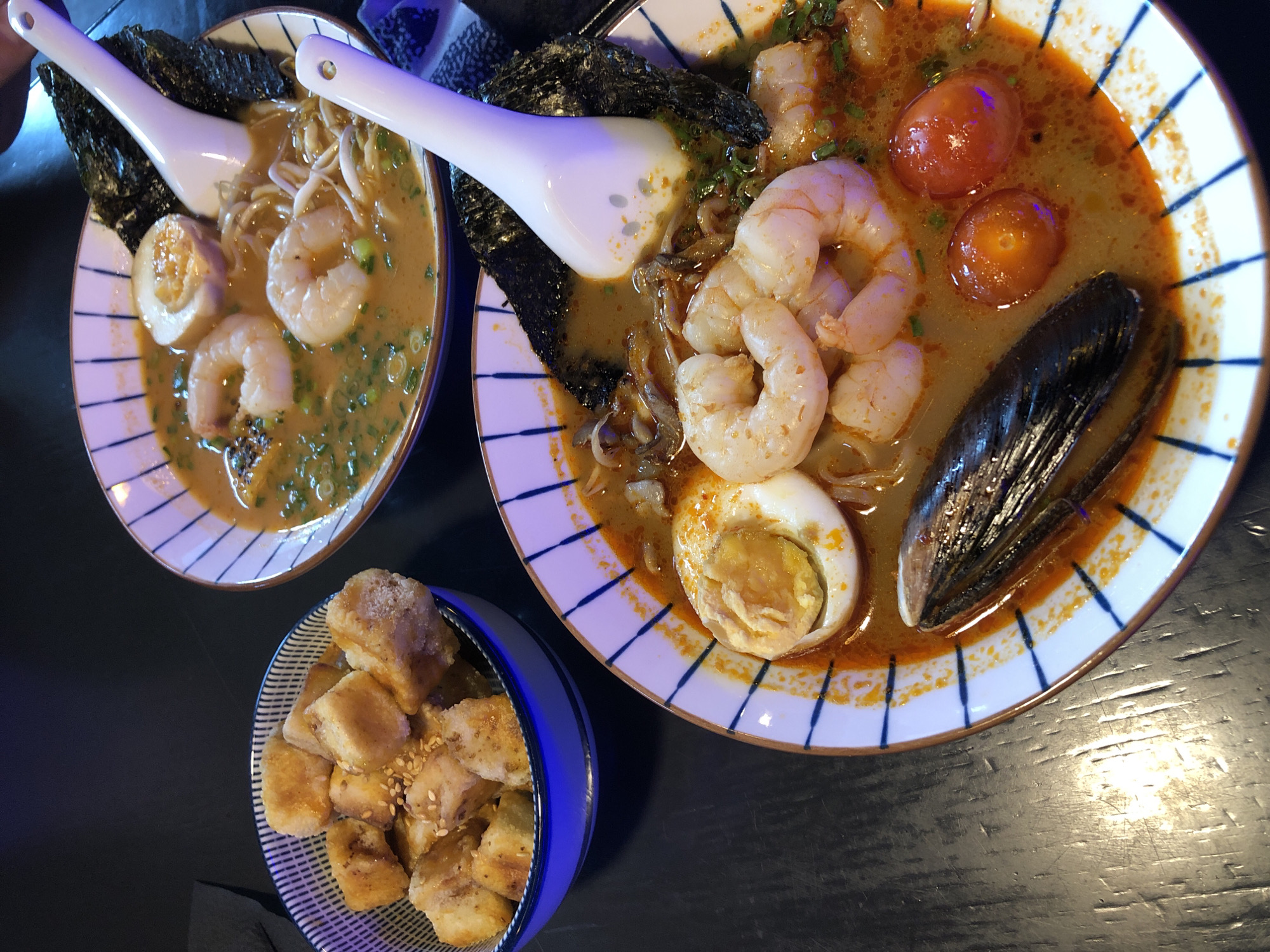 Two bowls of ramen soup are presented on a dark, reflective surface, possibly a table. One bowl is in the foreground and the other is slightly further back and to the right. Both bowls are white with blue striped rims.
The foreground bowl contains a reddish-orange broth, noodles, three large shrimp, a hard-boiled egg cut in half, a mussel shell, two small cherry tomatoes, and chopped green onions. A large white spoon rests in the broth, partially obscuring some of the ingredients. A piece of dark seaweed is visible on the right side of the bowl.
The background bowl contains a lighter orange broth, noodles, two shrimp, a hard-boiled egg cut in half, and chopped green onions. A large white spoon is also in this bowl, resting against a piece of dark seaweed.
To the left of the bowls, a smaller blue and white striped bowl is filled with golden-brown, bite-sized pieces of food, possibly fried tofu or chicken, sprinkled with sesame seeds. The overall lighting suggests an indoor setting, possibly a restaurant.
There are no people visible in the image. The image was taken from a slightly elevated angle, looking down at the food. The background is dark and blurred, with hints of purple and blue light reflecting on the surface. The scene appears to be a close-up, overhead shot of a meal. It is not possible to determine the location in Izhevsk, Russia, from the visual information provided.