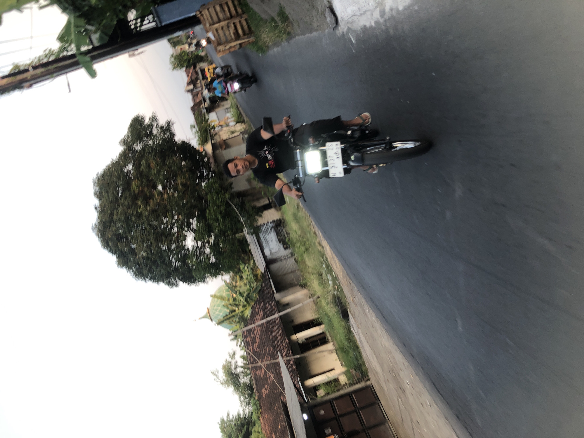 In Glagah Arum, Indonesia, a man is riding a black motorcycle down a paved road. He is wearing a black t-shirt and shorts, with white sandals. The motorcycle has a white license plate. In the background, there are houses with red tile roofs, a large tree, and a green domed building, possibly a mosque. Another person is riding a motorcycle further down the road, and another person is visible in the distance. A utility pole with wires stands on the right side of the road, next to stacked wooden pallets. The sky is overcast.