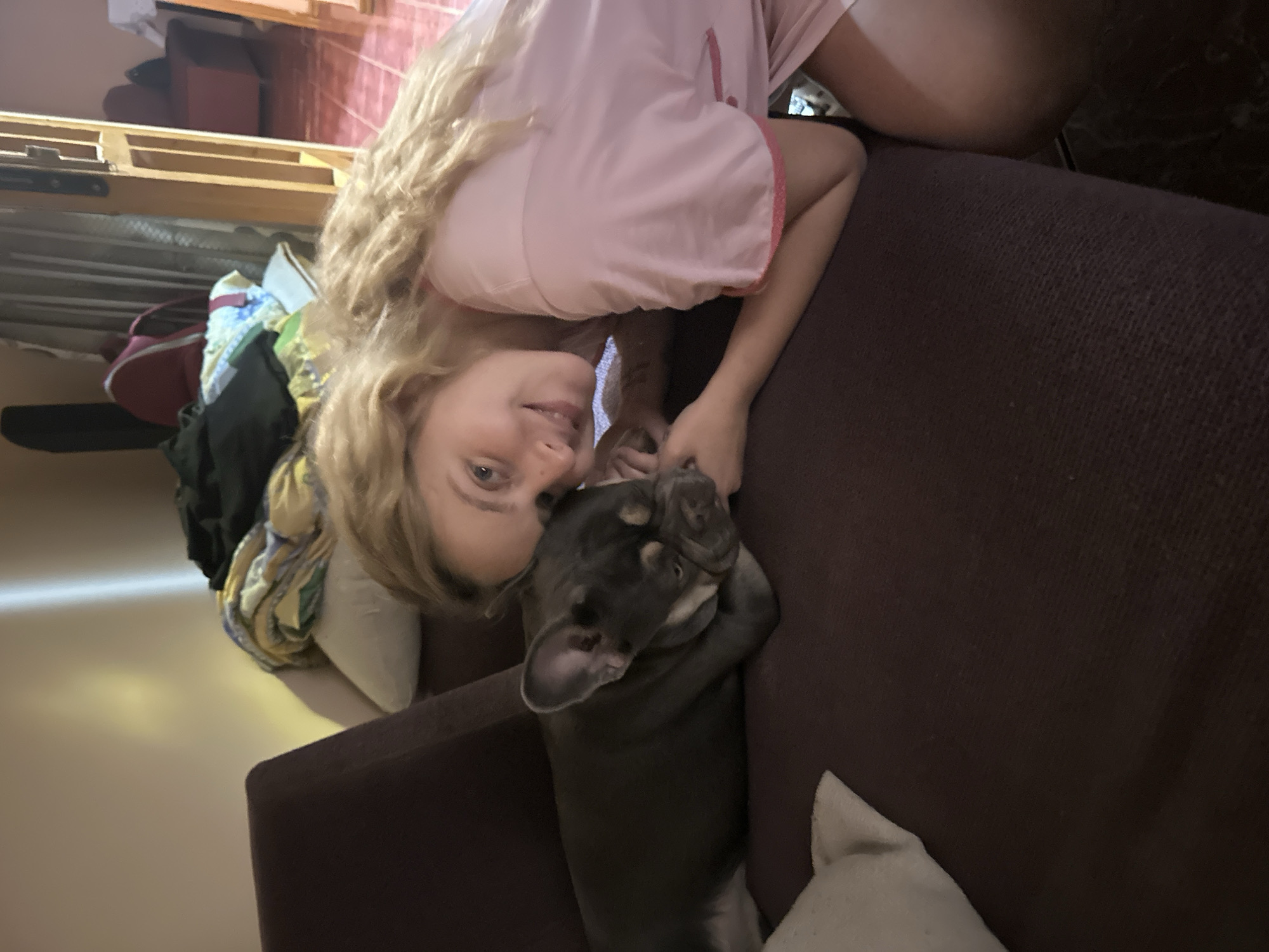 A young woman with long blonde hair, wearing a light pink pajama top, is lying on a dark brown upholstered couch with a gray French bulldog. The woman is smiling at the camera and holding the dog's paw. The scene is indoors, with a messy area in the background. The location is Unknown, Unknown.