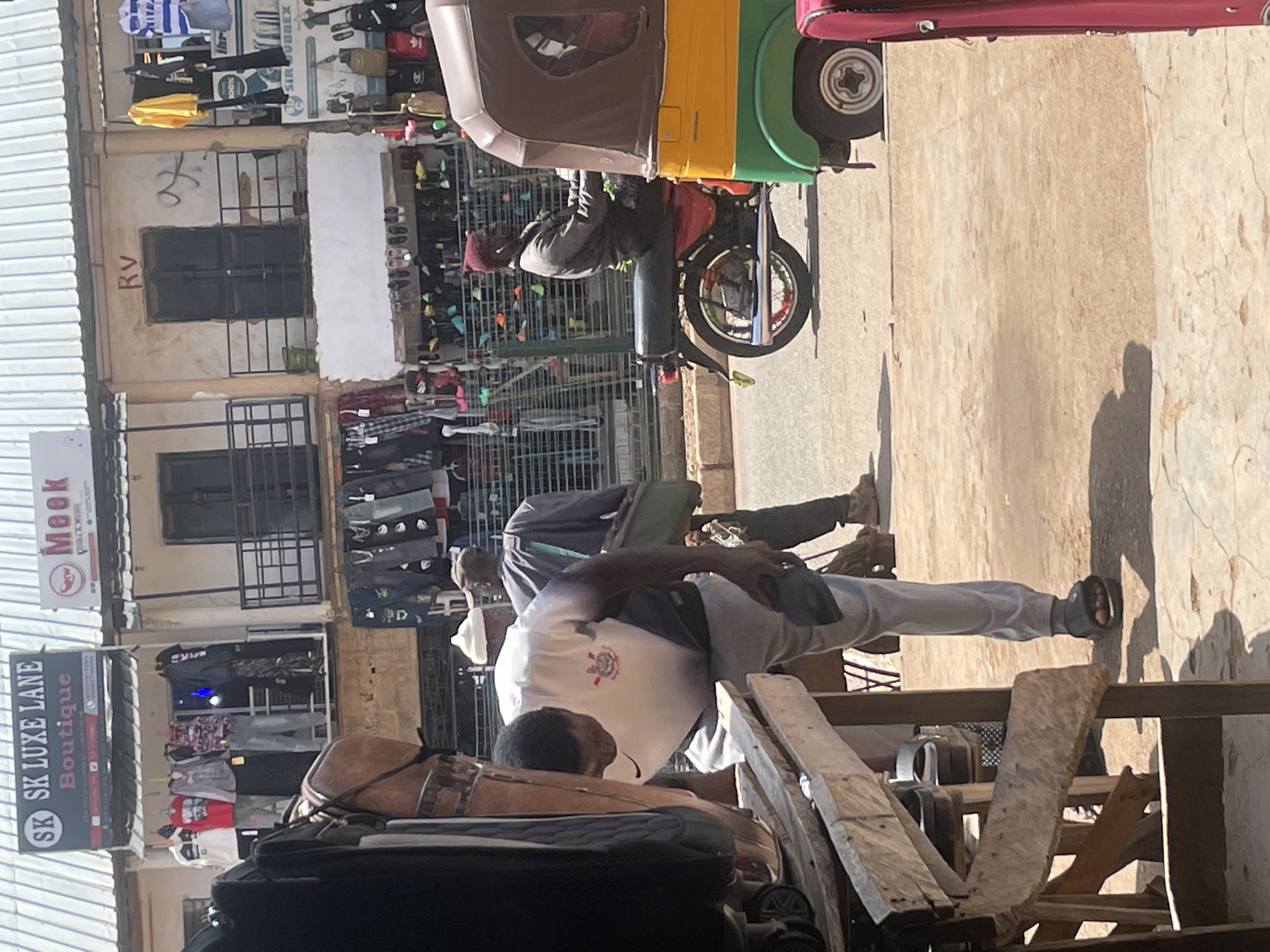 In Bukuru, Nigeria, a street market scene unfolds under a bright, overcast sky. The foreground is dominated by a rough wooden stall, with a person in a white and grey t-shirt and grey sweatpants leaning over it, examining something out of frame. This individual is wearing black sandals. Behind them, two other men are visible: one in a dark purple shirt and trousers carrying a green bag, and another wearing a white cap and grey attire. To the right, a three-wheeled auto-rickshaw, colored yellow, green, and brown, is parked. Its driver, a man in a grey jacket and dark cap, is seated on a red motorcycle that is attached to the auto-rickshaw. Further in the background, shops line the street. One shop displays a sign for "SK Luxe Lane Boutique" with clothing items hanging outside. Another sign reads "Meek." Various garments and footwear are displayed on racks and shelves along the storefronts. The ground is a mixture of dirt and paved surfaces, with shadows cast from the buildings and stalls. The overall impression is of a bustling commercial area in an urban environment.