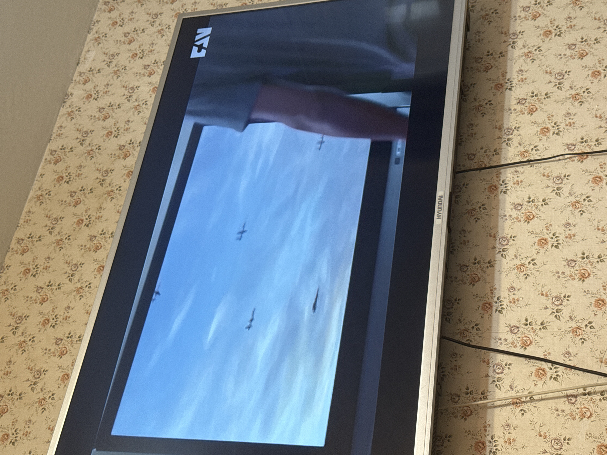 The image shows a Hyundai television displaying a video of fighter jets flying in formation against a blue sky. A person's arm is visible at the top of the screen. The television is mounted on a wall covered in floral wallpaper. There is no indication of this photo being taken in Mytishchi, Russia.