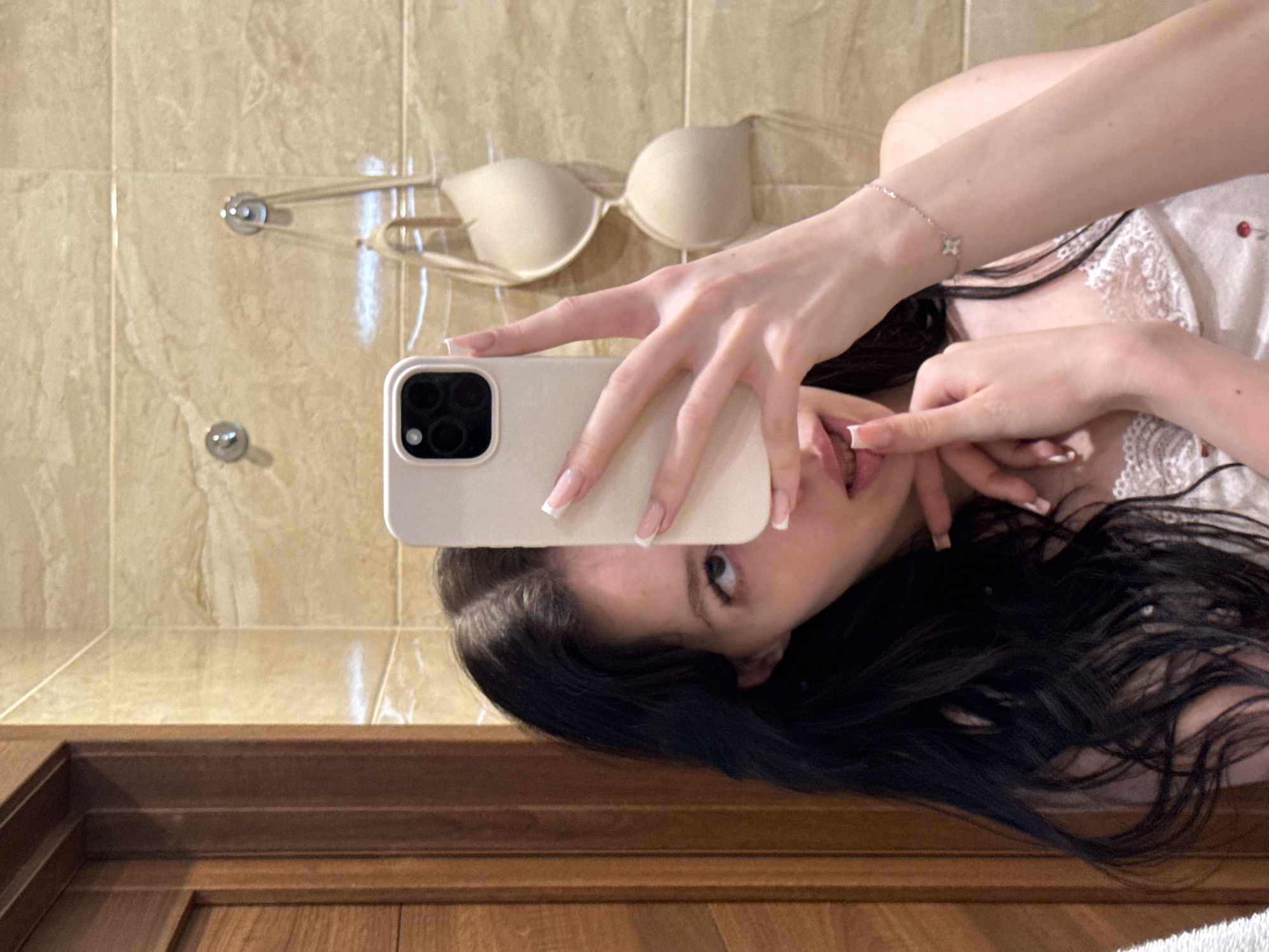 A young woman with long, dark hair takes a selfie in a bathroom with beige tiled walls. She is wearing a light-colored top with lace trim. Her left hand is holding a phone to take the picture, while her right index finger is near her mouth. A beige bra is hanging on hooks on the wall behind her. The environment appears to be a private residence in the city of Khimki, Russia.
