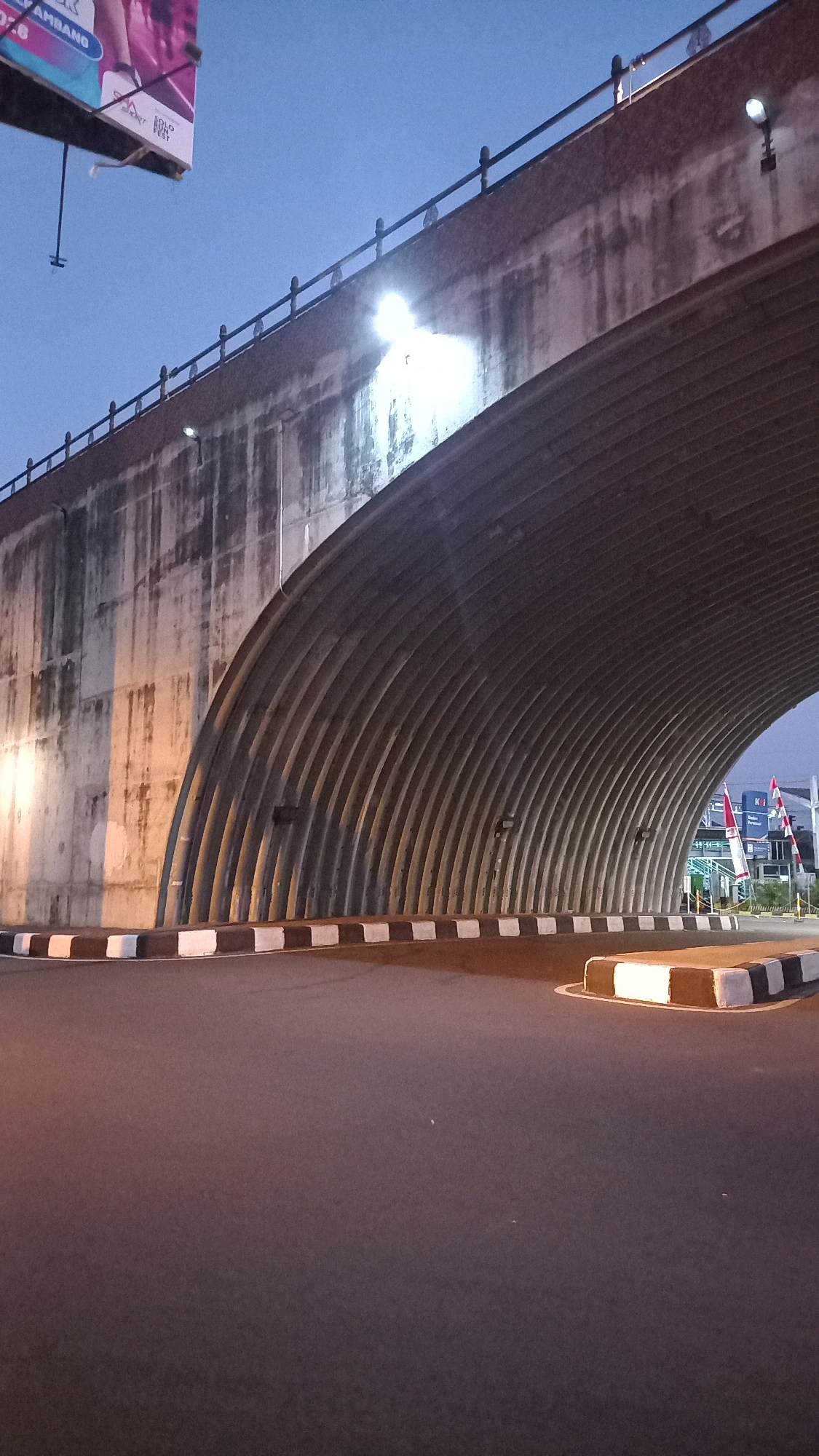 This image depicts the underside of a concrete overpass, likely part of a road infrastructure in the city of Surakarta, Indonesia. The overpass features a distinctive ribbed concrete ceiling, forming a tunnel-like appearance. The concrete walls of the overpass are stained and weathered, with artificial lighting illuminating the scene. The ground level shows a paved road with a clear boundary marked by alternating black and white blocks. In the background, hints of urban life are visible, including buildings, flags, and what appears to be a billboard overhead on the left side of the frame. The sky is a deep twilight blue, suggesting the image was taken during either dawn or dusk. There are no people or vehicles visible in the immediate foreground, creating a sense of stillness and emptiness within the urban landscape.