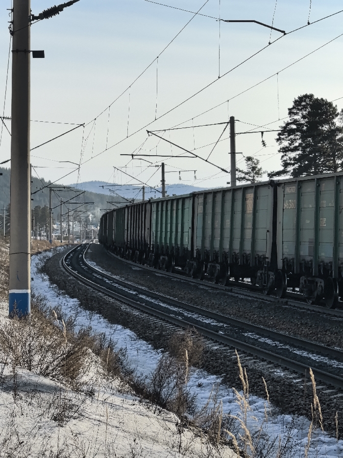 A freight train, composed of numerous light green/teal open-top railcars, travels along a set of two railway tracks curving towards the left in the city of Atamanovka, Russia. The tracks are ballasted and lined with patchy snow cover and sparse, dry vegetation in the foreground. Overhead catenary lines, supported by multiple grey utility poles, span above the tracks. The closest pole in the left foreground exhibits a blue band painted around its base. In the background, rolling, snow-dusted hills are visible beneath a pale blue sky, with scattered evergreen trees along the right side of the tracks and faint distant structures to the left. No individuals are present. Visible markings on a railcar include "РЖД" and "644 3160".