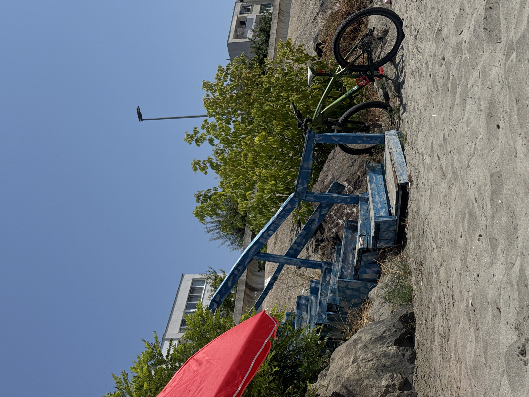 The image captures a serene outdoor scene on a sunny day in Pomorie, Bulgaria. In the foreground, a sandy beach stretches out, with fine-grained sand showing subtle ripple patterns. To the left, a large, weathered grey rock formation anchors the scene. Partially obscuring the view is a vibrant red beach umbrella, its fabric taut against the clear blue sky.
A set of weathered, blue-painted wooden stairs ascends from the beach, its paint peeling to reveal a patterned texture beneath, suggesting it has seen much use. These stairs are flanked by rocky terrain and a dense thicket of green foliage.
Resting on the sand to the right of the stairs is a green mountain bike. Its tires are slightly deflated, and a red water bottle cage is visible on the frame. The bike is propped up by its kickstand. In the background, a modern street lamp stands tall against the sky, and further back, a multi-story white building with a balcony is visible, nestled amongst lush green trees. Another building, appearing more residential with visible windows and a light-colored facade, is partially visible on the right side of the frame. The overall impression is one of a quiet, possibly tourist-frequented spot, enjoying the summer weather.