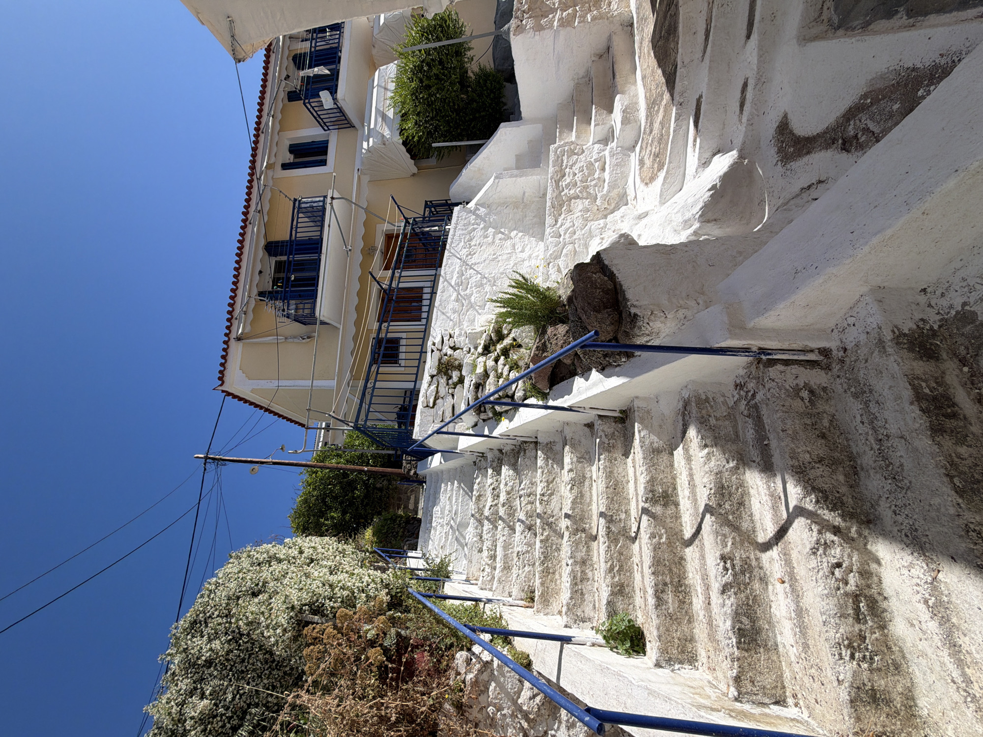 The image captures a street scene in Poros, Greece. A multi-story building with a yellow facade, red-tiled roof, and blue balconies is on the left. To its right, a whitewashed stone stairway with a blue metal railing ascends along a white stone wall. The sky is clear and blue. Vegetation, including trees and flowering shrubs, is also visible. An overhead electrical line crosses the upper part of the scene.