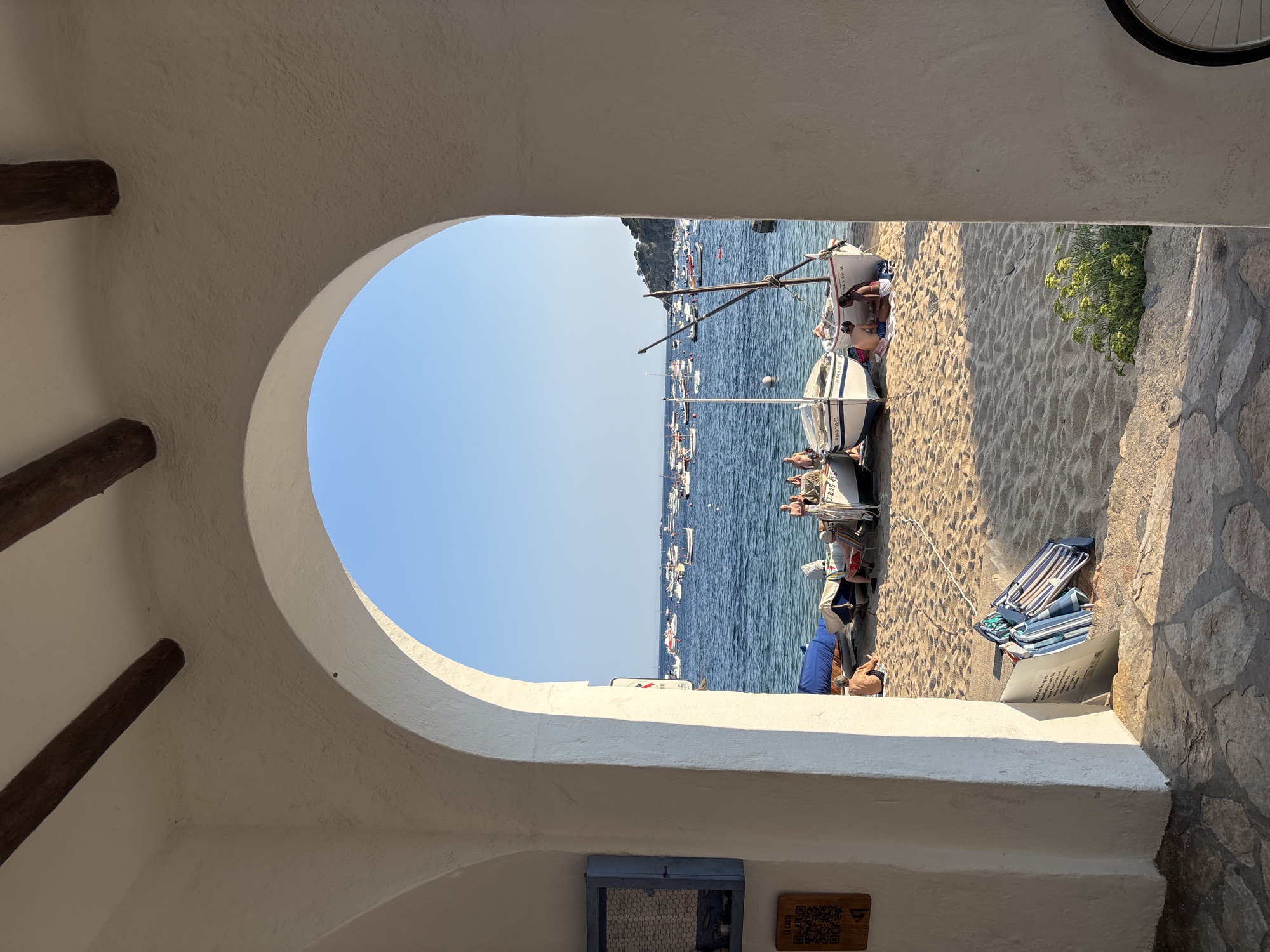 The image captures a serene beach scene in Calella De Palafrugell, Spain, viewed through a white arched passageway. Sunlight streams through the arch, illuminating a sandy beach dotted with several small, white fishing boats pulled ashore. Some of the boats are covered with blue tarps. A few people are visible on the beach, some sitting near the boats and others further back, appearing to be relaxing or sunbathing. Beyond the boats, the calm, blue sea stretches to the horizon, filled with numerous other boats of varying sizes, suggesting a busy harbor or bay. The sky is a clear, bright blue, indicating pleasant weather. In the foreground, within the archway, a portion of a textured white wall and a stone pathway are visible, along with a stack of folded beach chairs and a leaning piece of paper. A blue cabinet with a wire mesh front is mounted on the wall to the left. The overall impression is one of a peaceful coastal village on a sunny day.